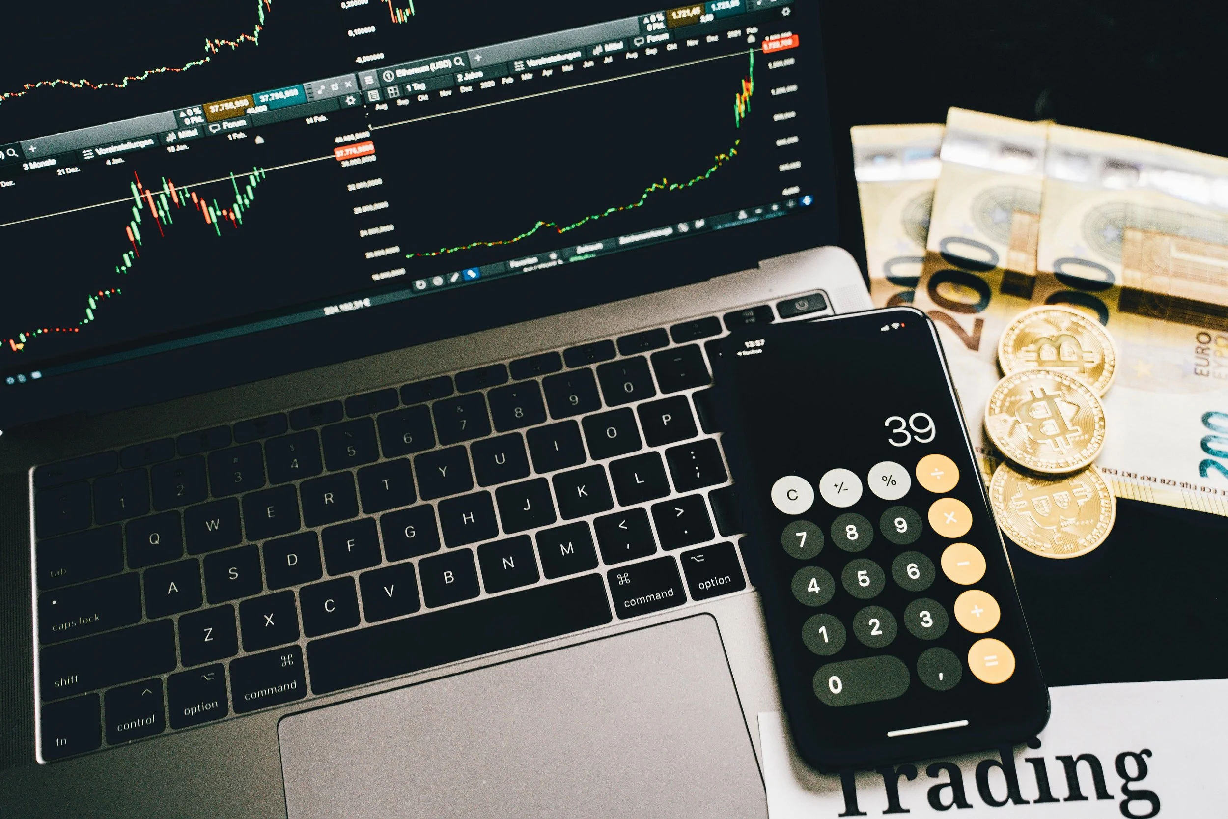 Pantalla de computadora portátil mostrando gráficos de mercado financiero, teléfono móvil con calculadora en pantalla y billetes de euro junto a monedas de bitcoin sobre una hoja con la palabra 'Trading'.