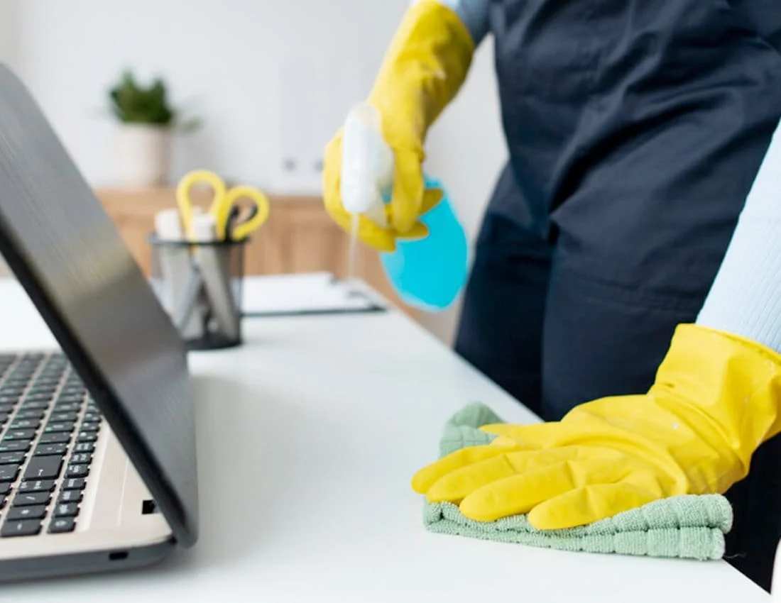 Person wearing yellow rubber gloves cleaning a white desk with a cloth and spray bottle in an office setting.