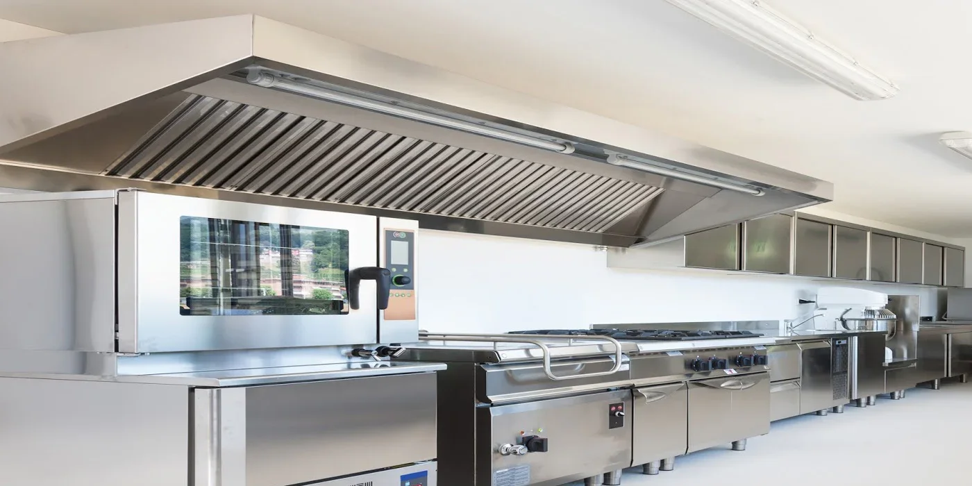 Industrial kitchen with stainless steel appliances, including a pizza oven and cooking range, under a large ventilation hood.
