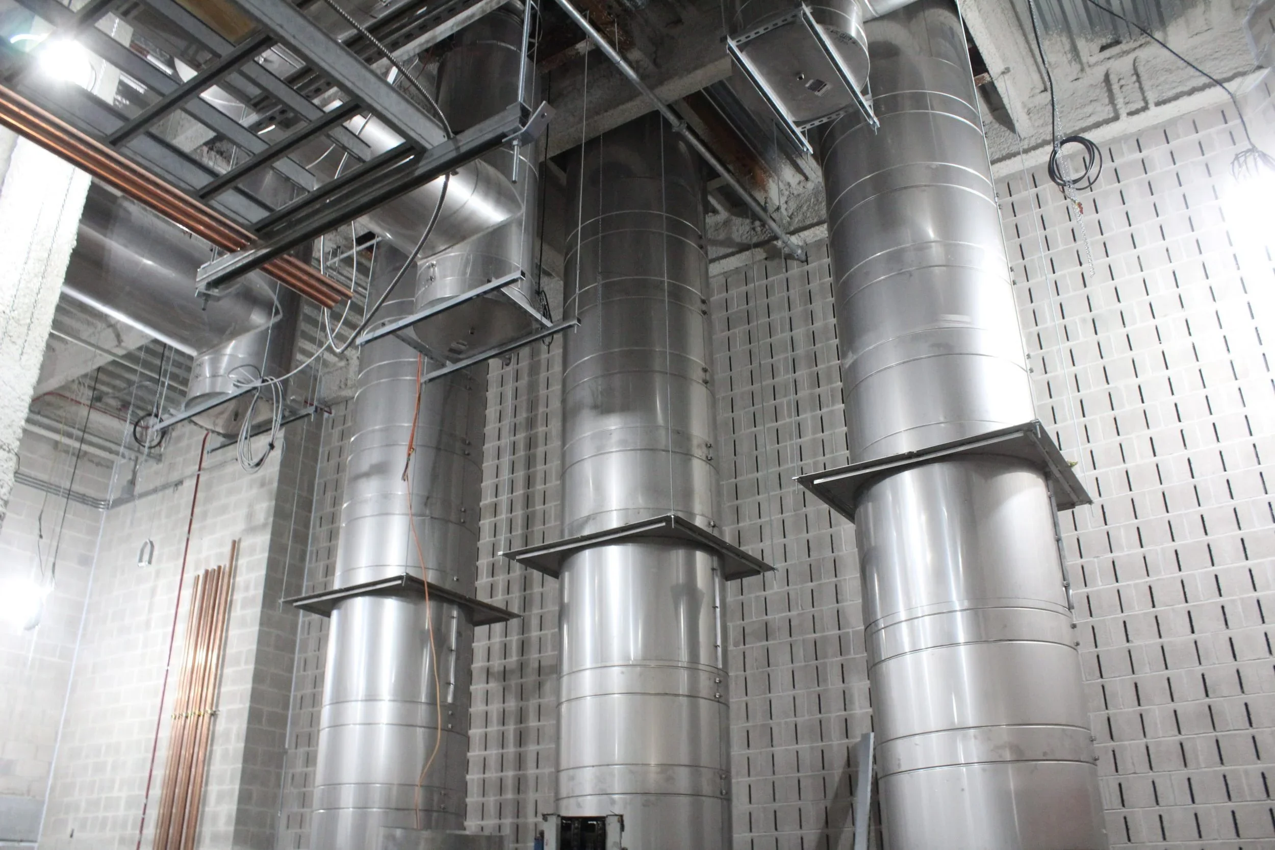 Construction site with large silver ventilation ducts running vertically against a partially finished brick wall, with exposed pipes and electrical wiring overhead.