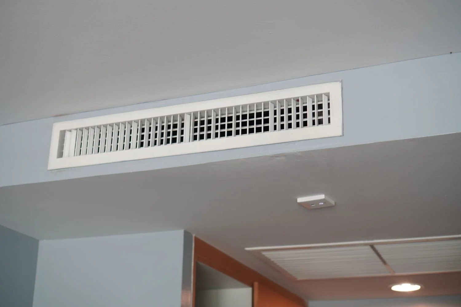 Ceiling air vent and ceiling-mounted device in a room with light gray walls and a wooden doorframe.