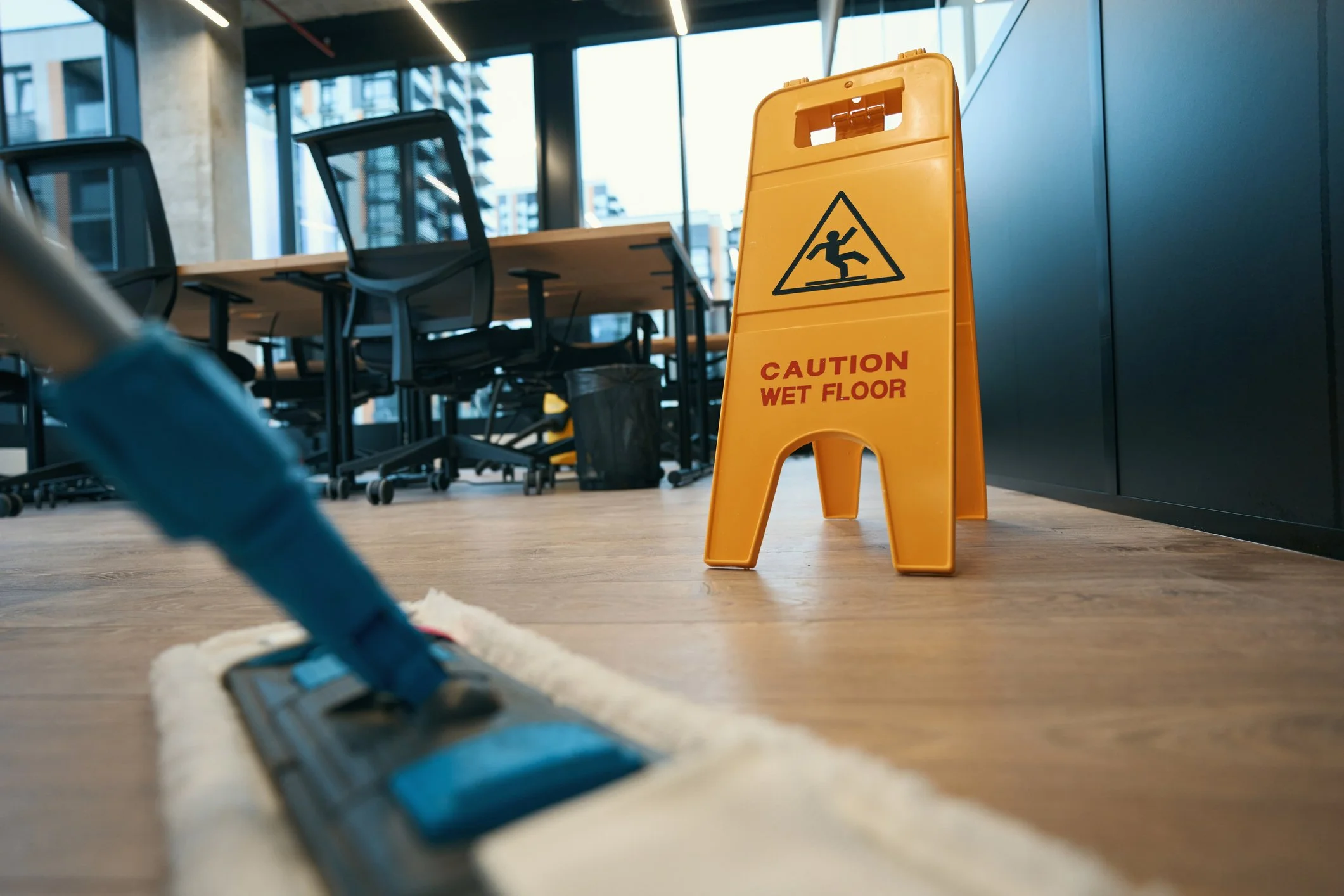 Close-up of a wet floor caution sign in front of a mop on the floor. The sign indicates a wet floor hazard in an indoor office or conference room with chairs and table set against large windows.