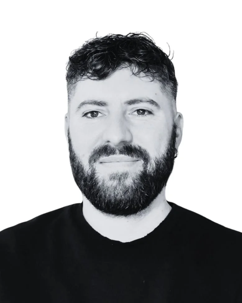 Black and white portrait of a man with dark, curly hair and a full beard, wearing a black shirt, against a plain background.
