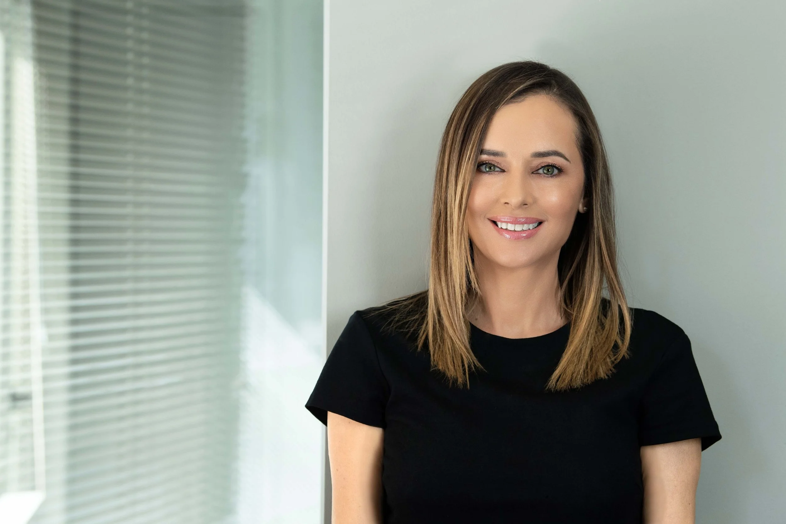 A woman with shoulder-length brown hair and green eyes, smiling, wearing a black shirt, standing indoors near a wall and frosted glass window.