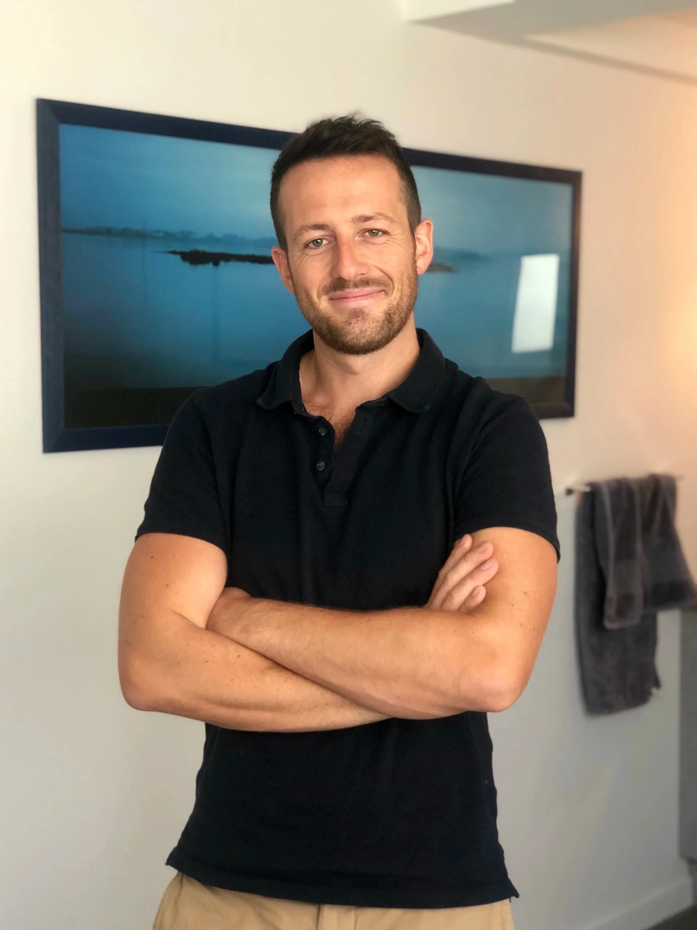 Homme souriant avec bras croisés, portant un polo noir, dans une pièce avec un tableau représentant un paysage aquatique derrière lui.
