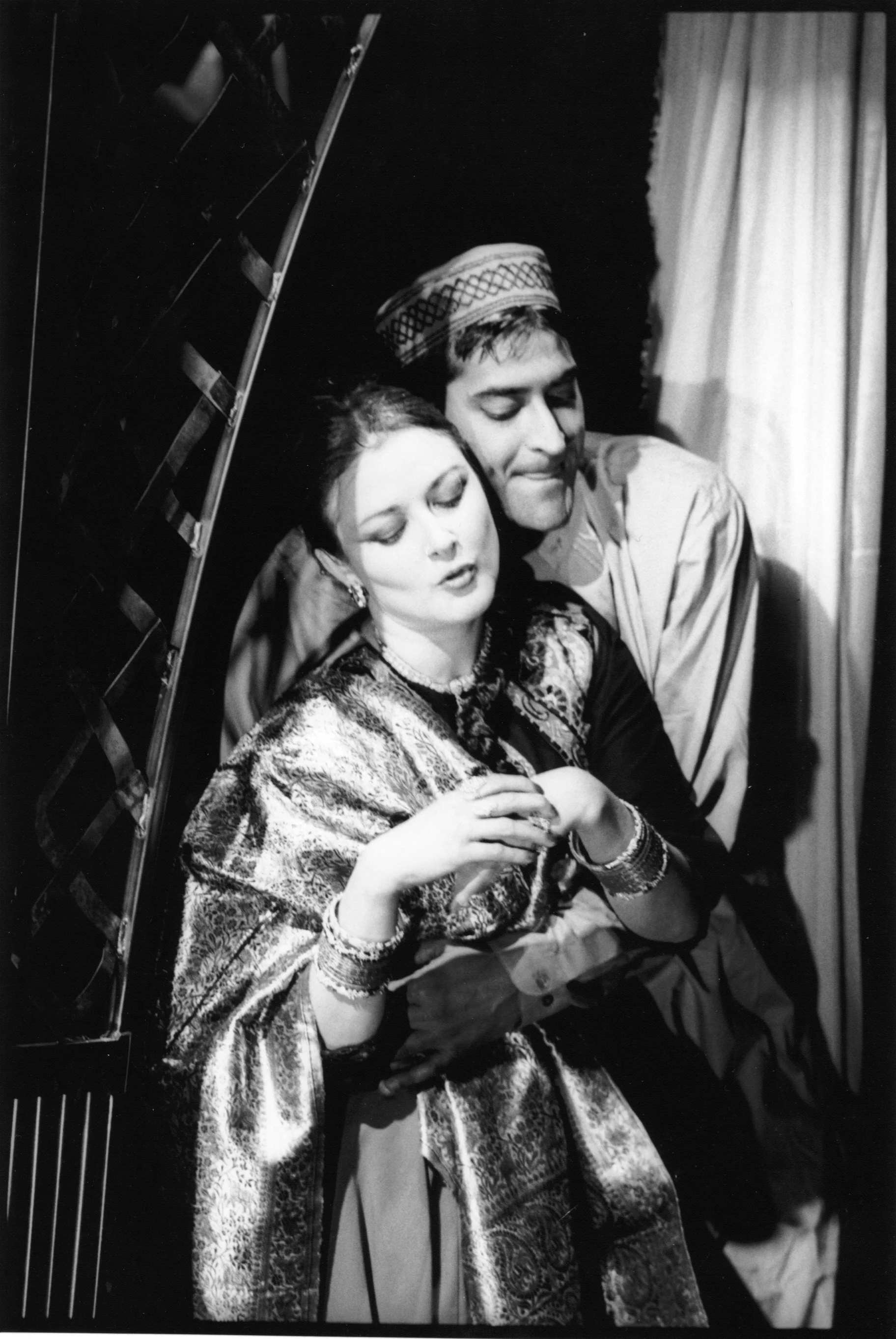 A black and white photo of a man and woman sitting closely together, with the woman in the foreground and the man behind her, both with their eyes closed. The woman is wearing a patterned dress and earrings, and the man is wearing traditional headwear and a shirt. They appear to be in an intimate or tender moment.