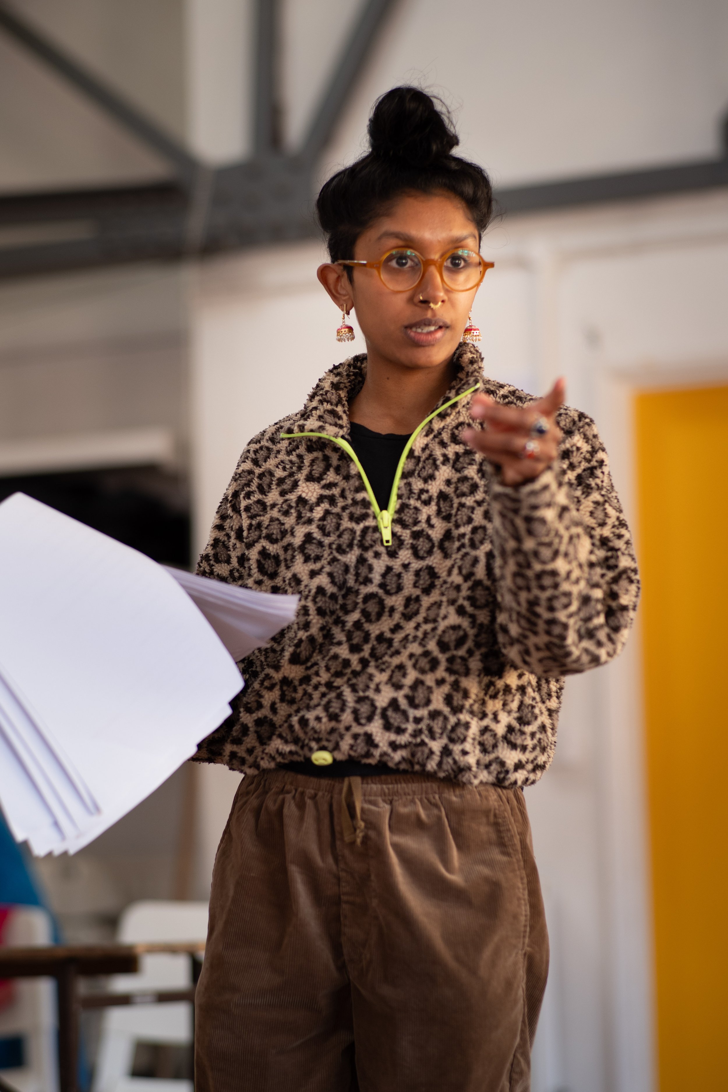 A person with glasses and earrings, wearing a leopard-print fleece jacket and brown pants, gesturing with their hand while speaking.