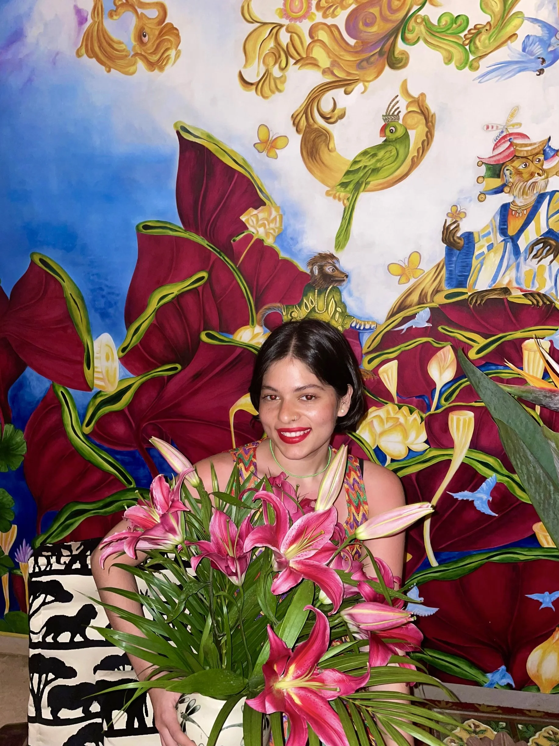 A woman with dark hair and a colorful dress smiling in front of a vibrant mural with large flowers, leaves, and cartoon animals, including a dog and a lion. She is holding a bouquet of pink lilies.