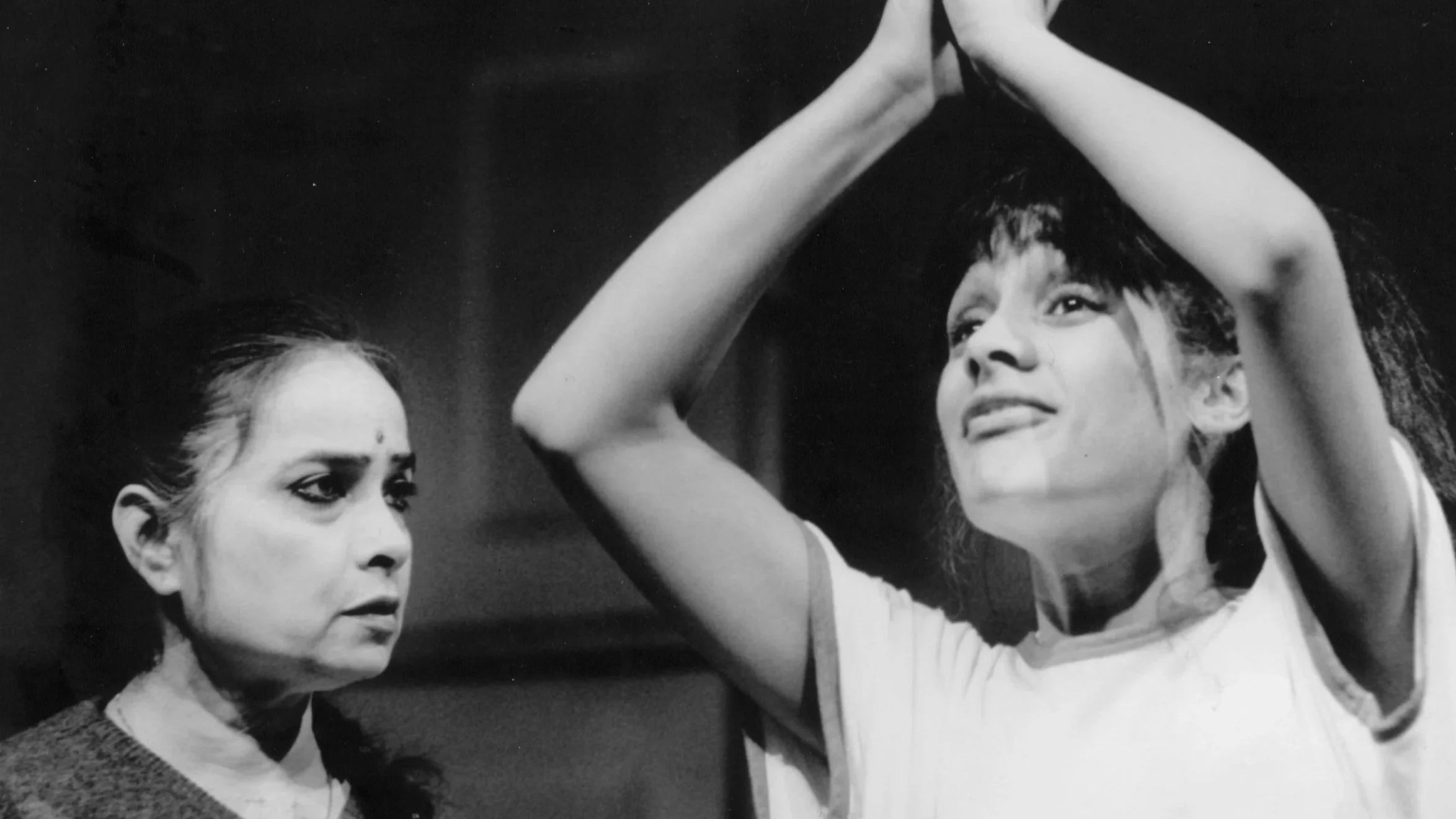 Two women with dark hair and serious expressions; one is looking to the side while the other is raising her arms with hands clasped above her head against a dark background.
