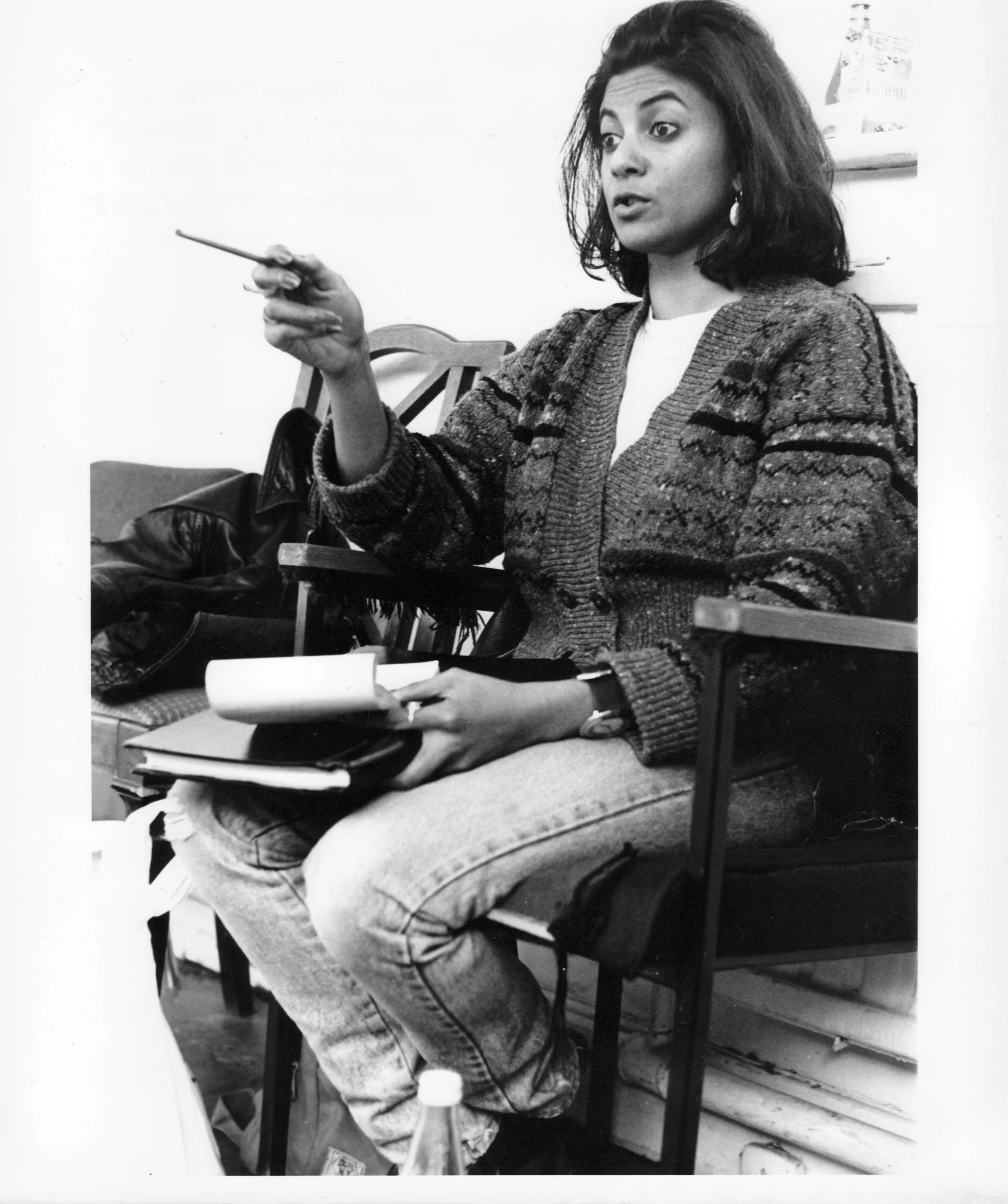 Black and white photo of a woman in casual clothing sitting on a chair, holding a notebook and pen, with papers on her lap, in a room with a white wall, some shelves, and bags on the floor.