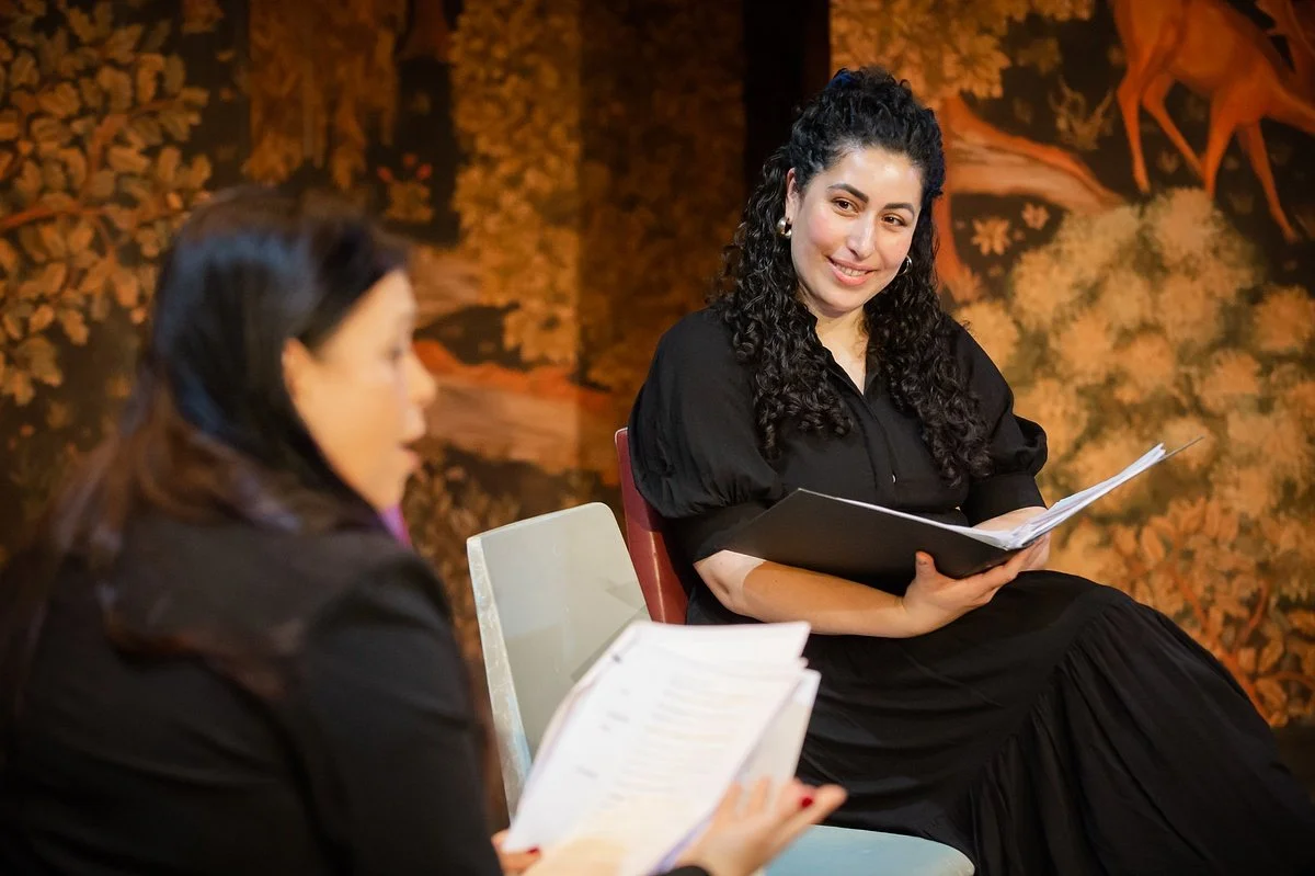 Two women sitting and talking, one with long dark hair and the other with curly hair, both holding papers, in a warmly lit room with autumn-themed decor.