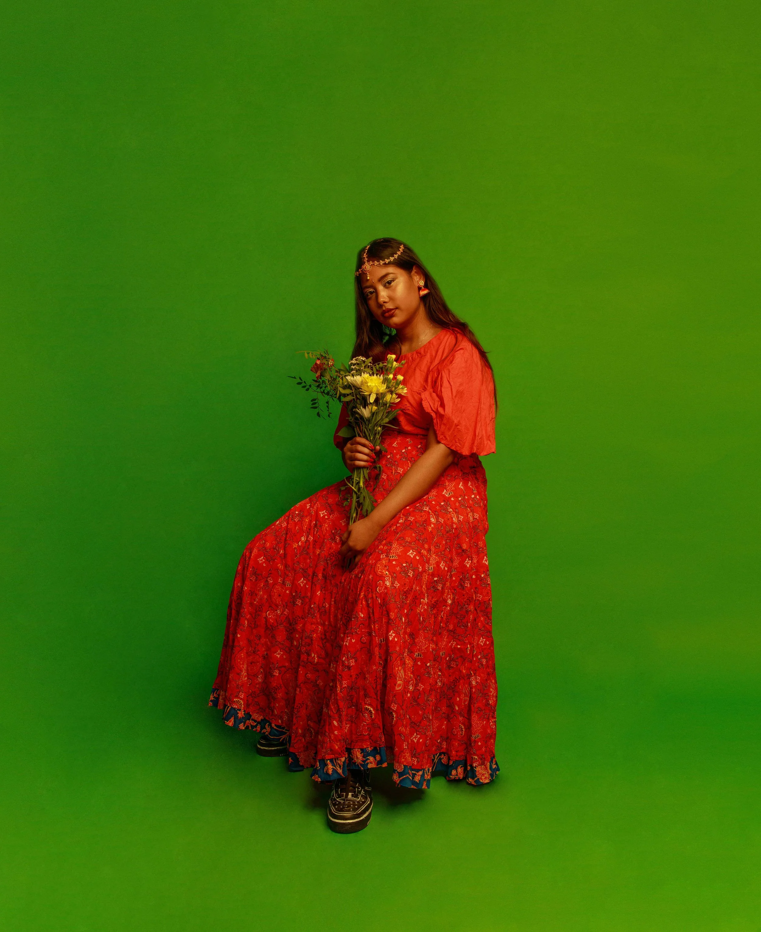 A woman in a red dress with floral patterns holding a bouquet of flowers, standing against a green backdrop, wearing jewelry including earrings and a headpiece.