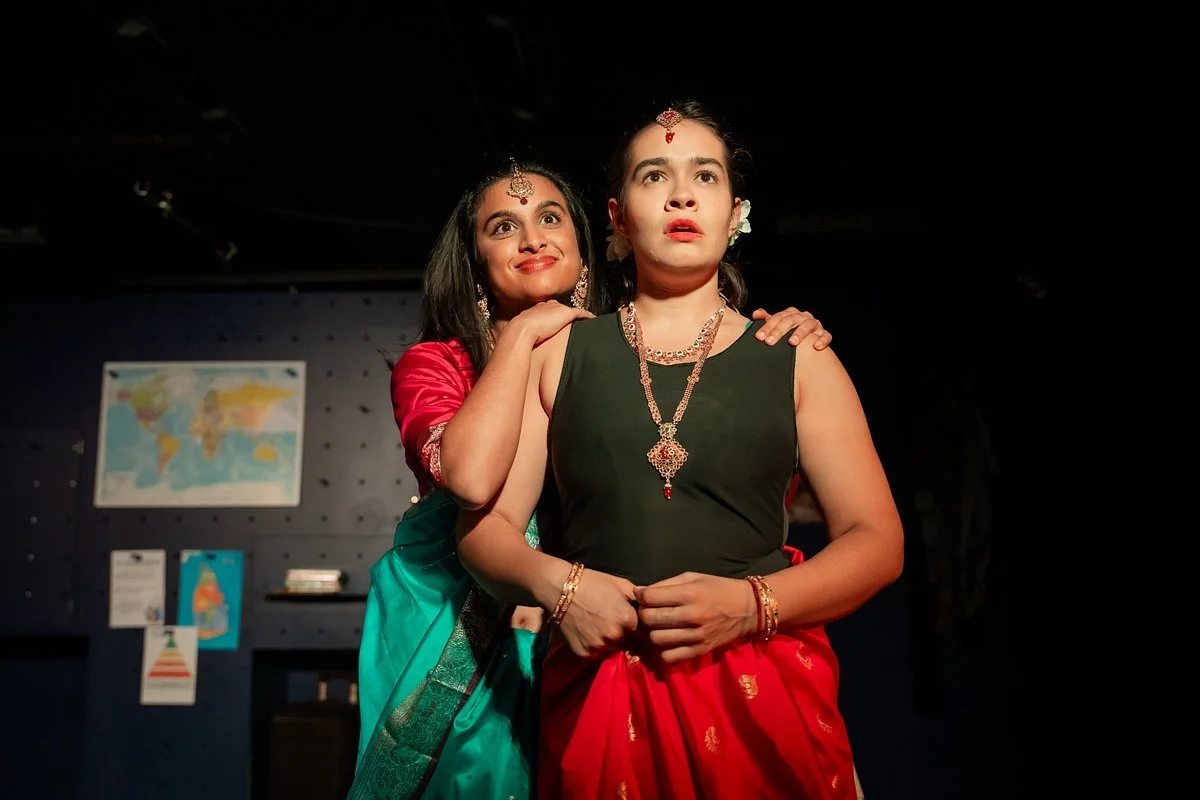 Two women dressed in traditional Indian attire stand closely together. One, smiling, places her hand on the other's shoulder. They wear jewelry and floral accessories in their hair, with a map and posters on a dark background behind them.