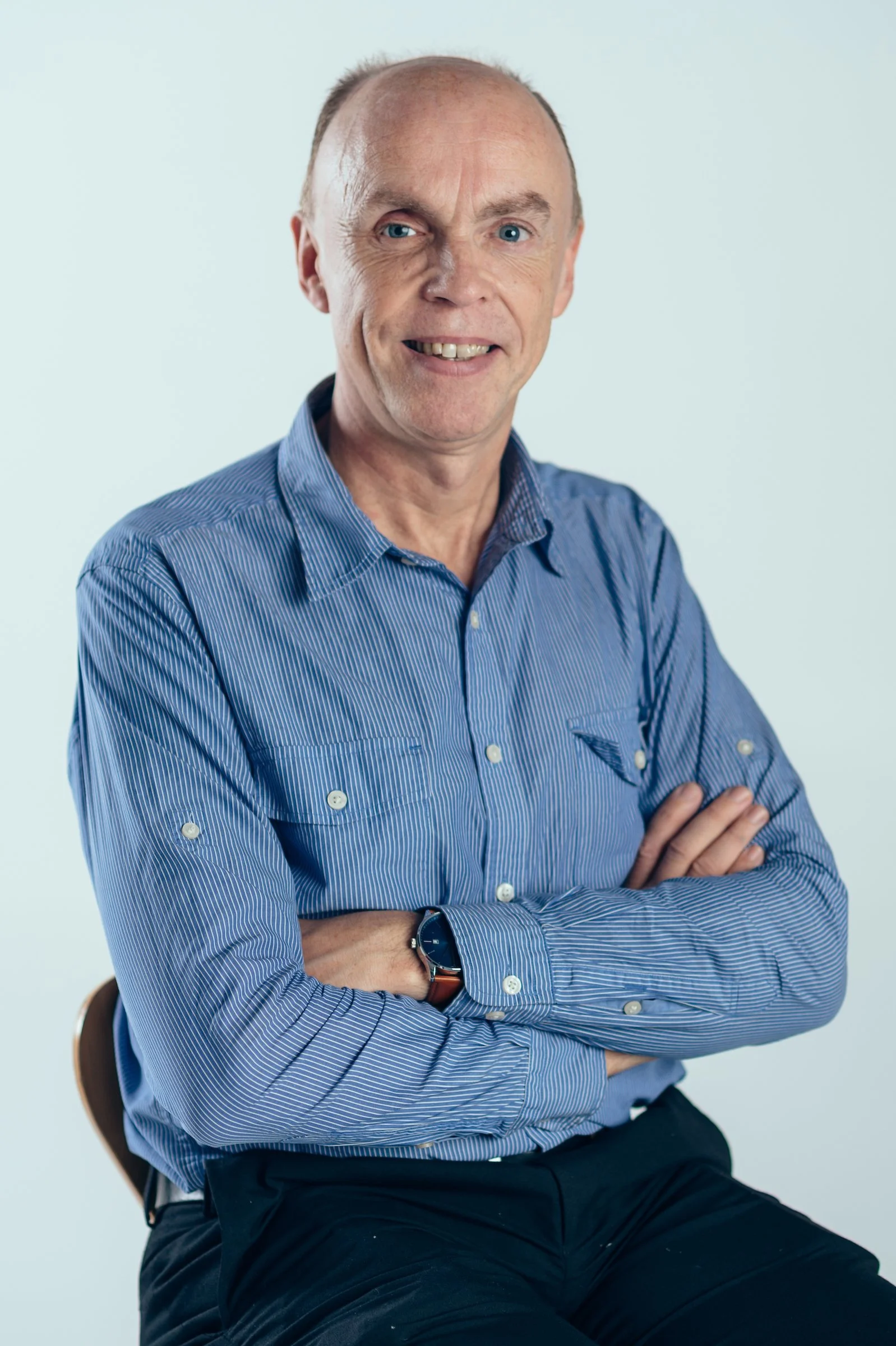 A middle-aged man with blue eyes, short hair, and a friendly smile, wearing a blue striped button-up shirt and sitting with crossed arms against a plain light background.