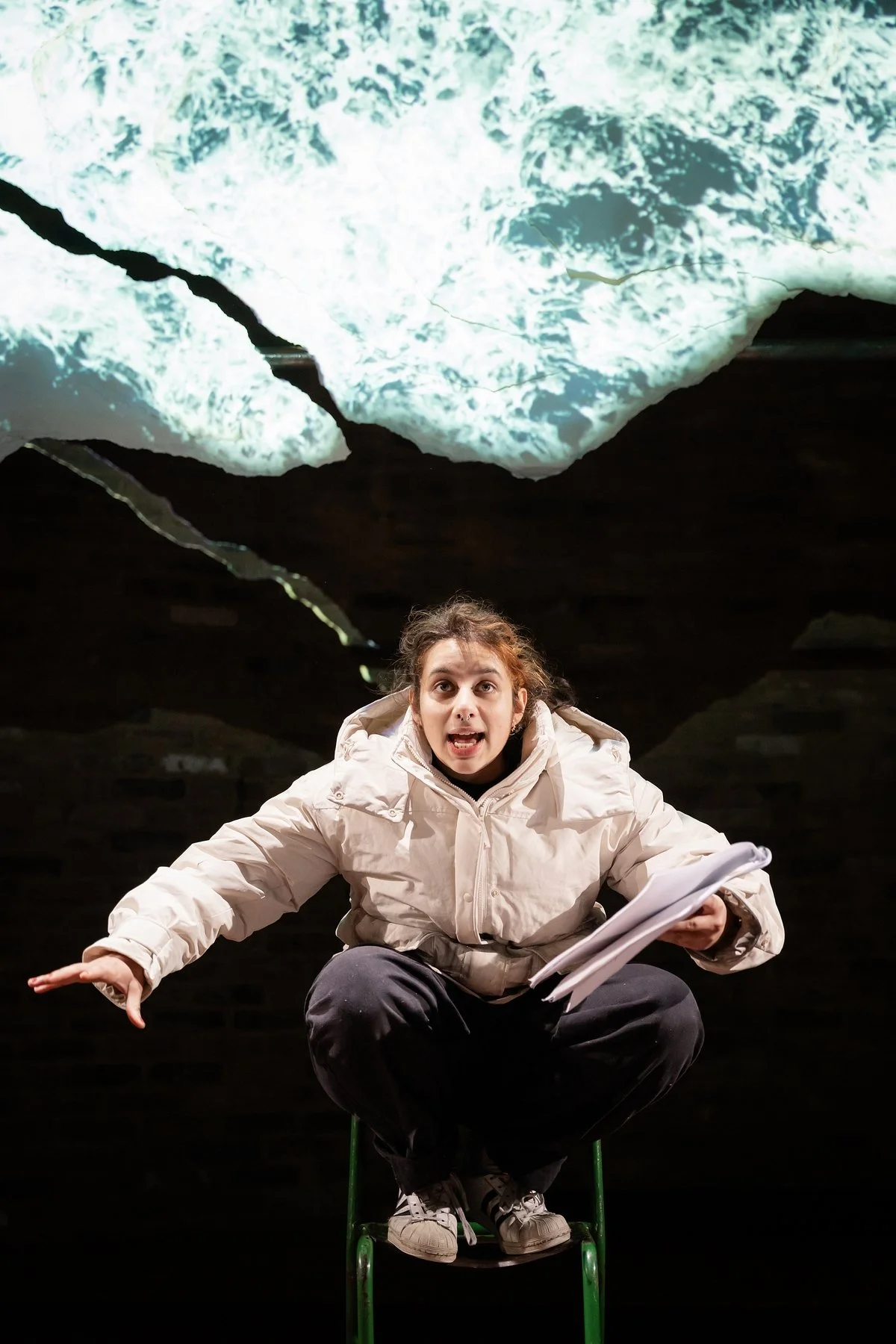 A woman crouching on a green chair, speaking passionately, in front of a dark background with an image of turbulent water projected above her.