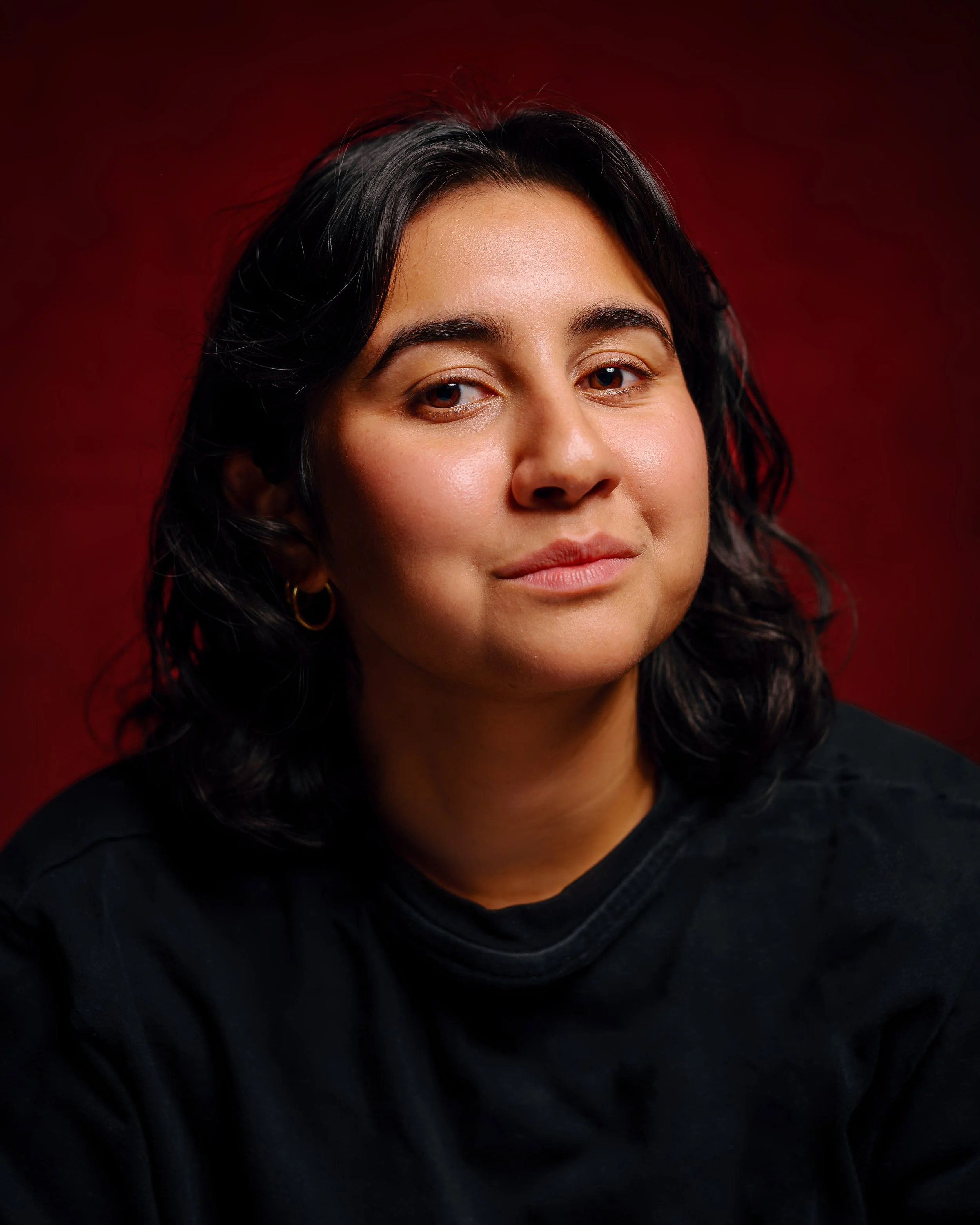 Close-up of a woman's face with black hair, wearing a black top, against a dark red background.