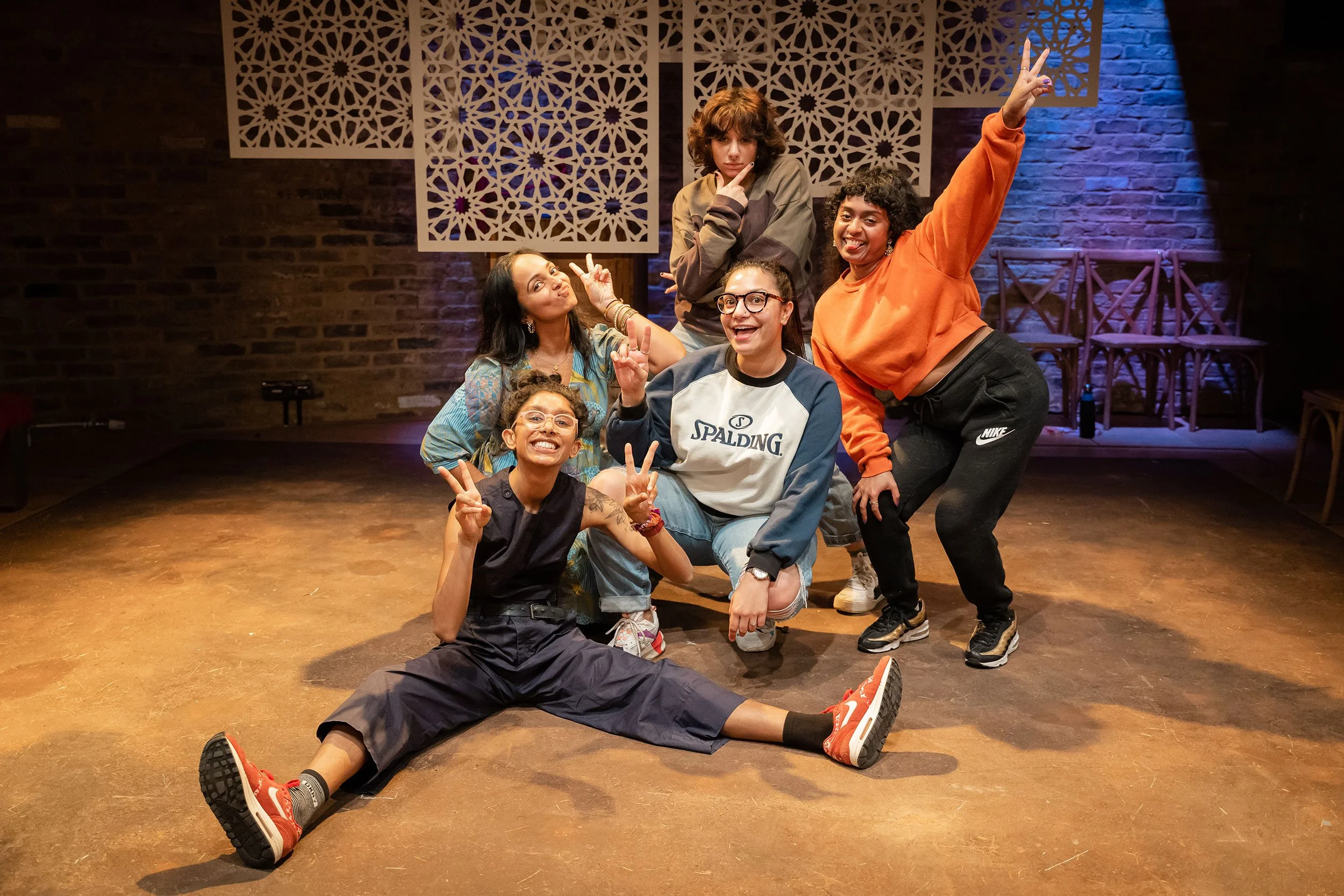 Group of five diverse young women posing playfully in a room with decorative wall panels and brick wall background. One woman is sitting on the floor with her legs extended, smiling and making peace signs. Others stand or crouch around her, smiling, making peace signs, and striking playful poses, with one woman raising her hand in a victory sign.