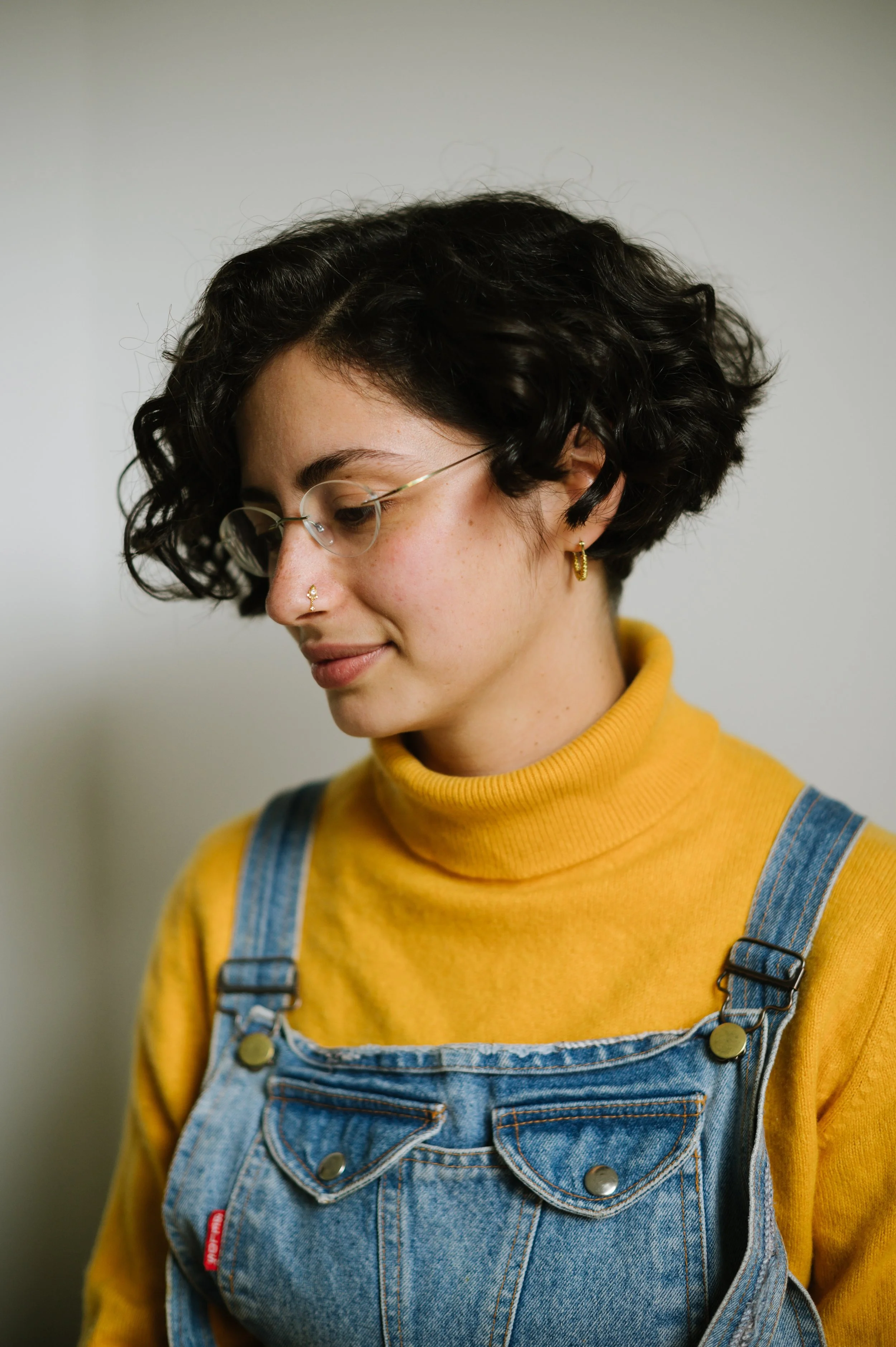 A young woman with dark curly hair wearing glasses, gold hoop earrings, and a yellow turtleneck sweater under blue denim overalls. She is facing slightly downward with a gentle smile, against a plain neutral background.