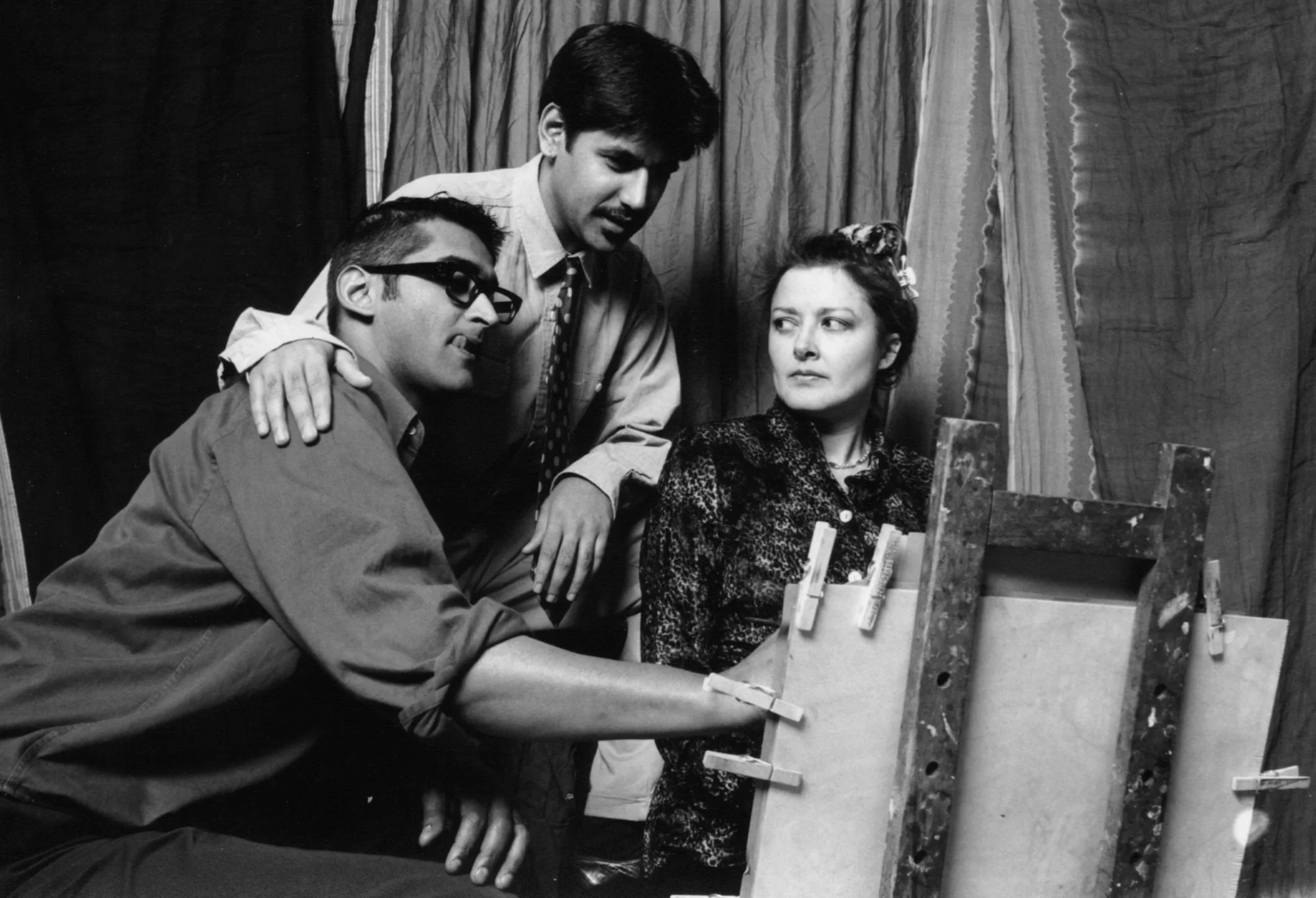 Black and white photo of two men and one woman engaged in a conversation around an art easel with canvases.