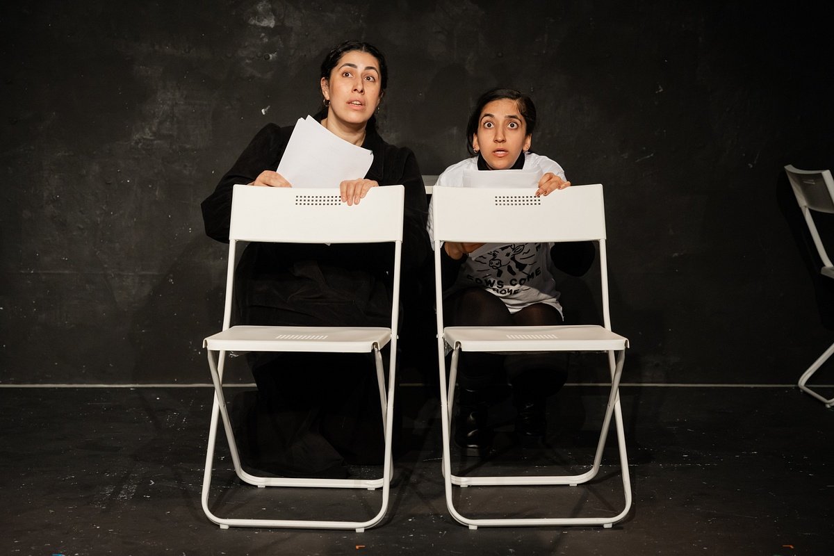 Two women sitting behind white chairs on a black stage, holding papers, with surprised or attentive expressions.