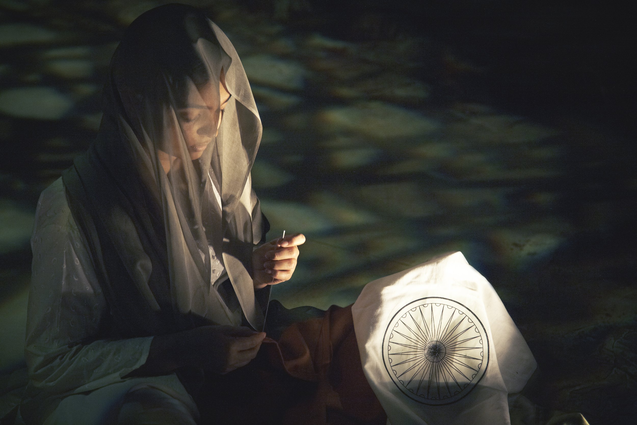 A person wearing a beige raincoat and a hood, looking down and holding a cigarette, sitting near water at night with greenish reflections.
