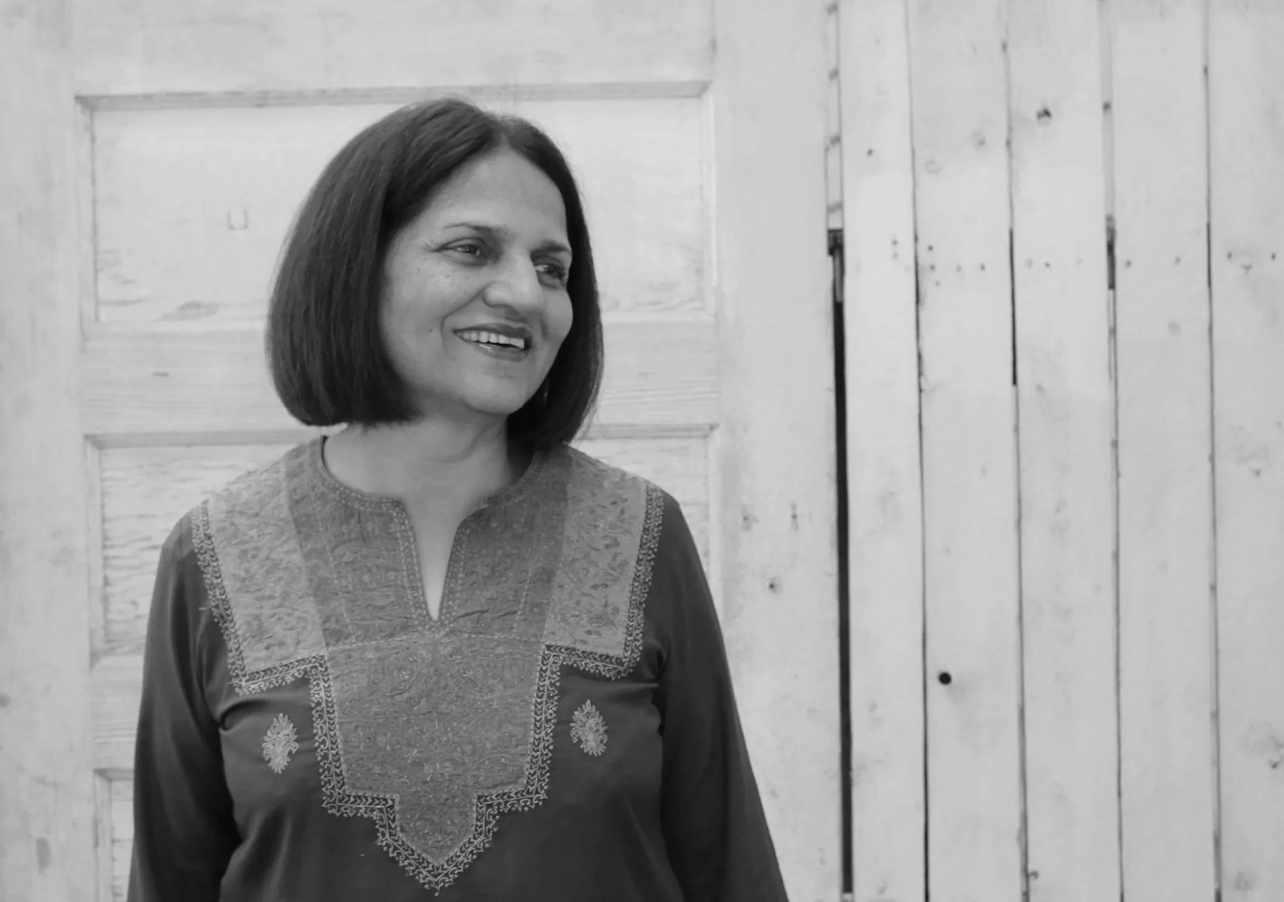 A woman with shoulder-length hair smiling, wearing a traditional embroidered top, standing in front of a wooden wall.
