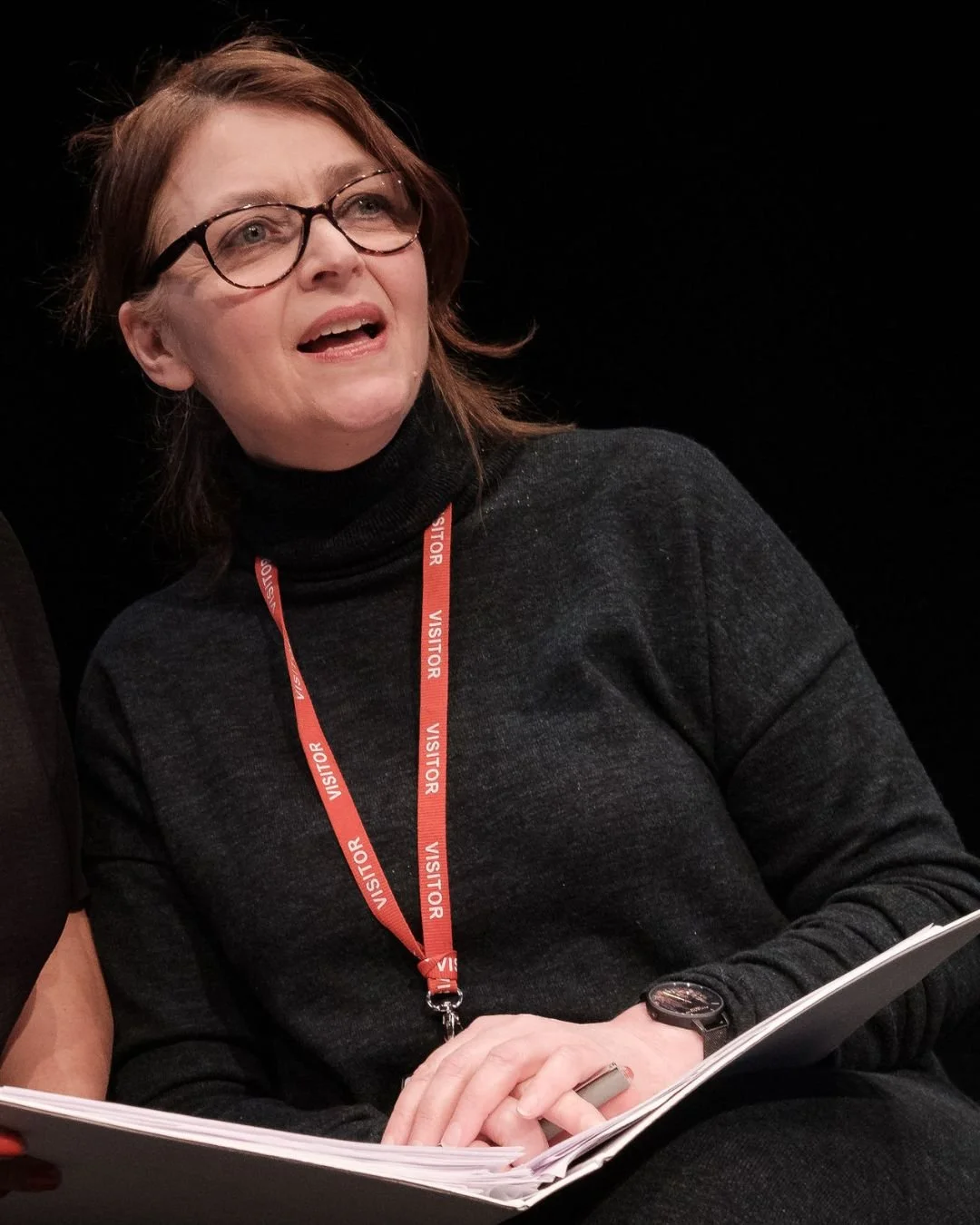 A woman with glasses and brown hair, wearing a black turtleneck sweater, holding papers, and wearing a red lanyard with the words 'VISITOR' repeatedly printed on it.