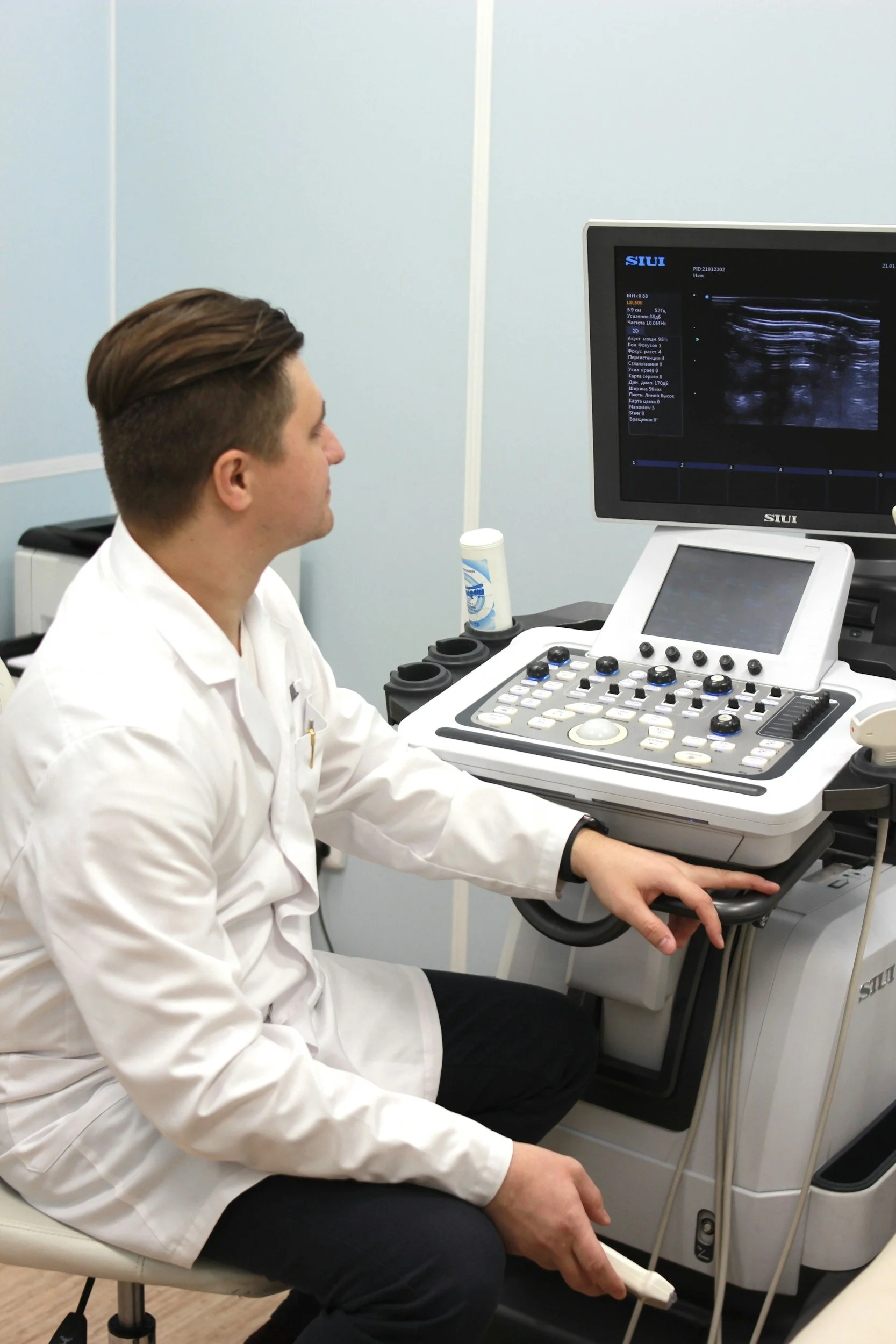 A man wearing a white lab coat sitting in front of an ultrasound machine, looking at the monitor displaying an ultrasound image.