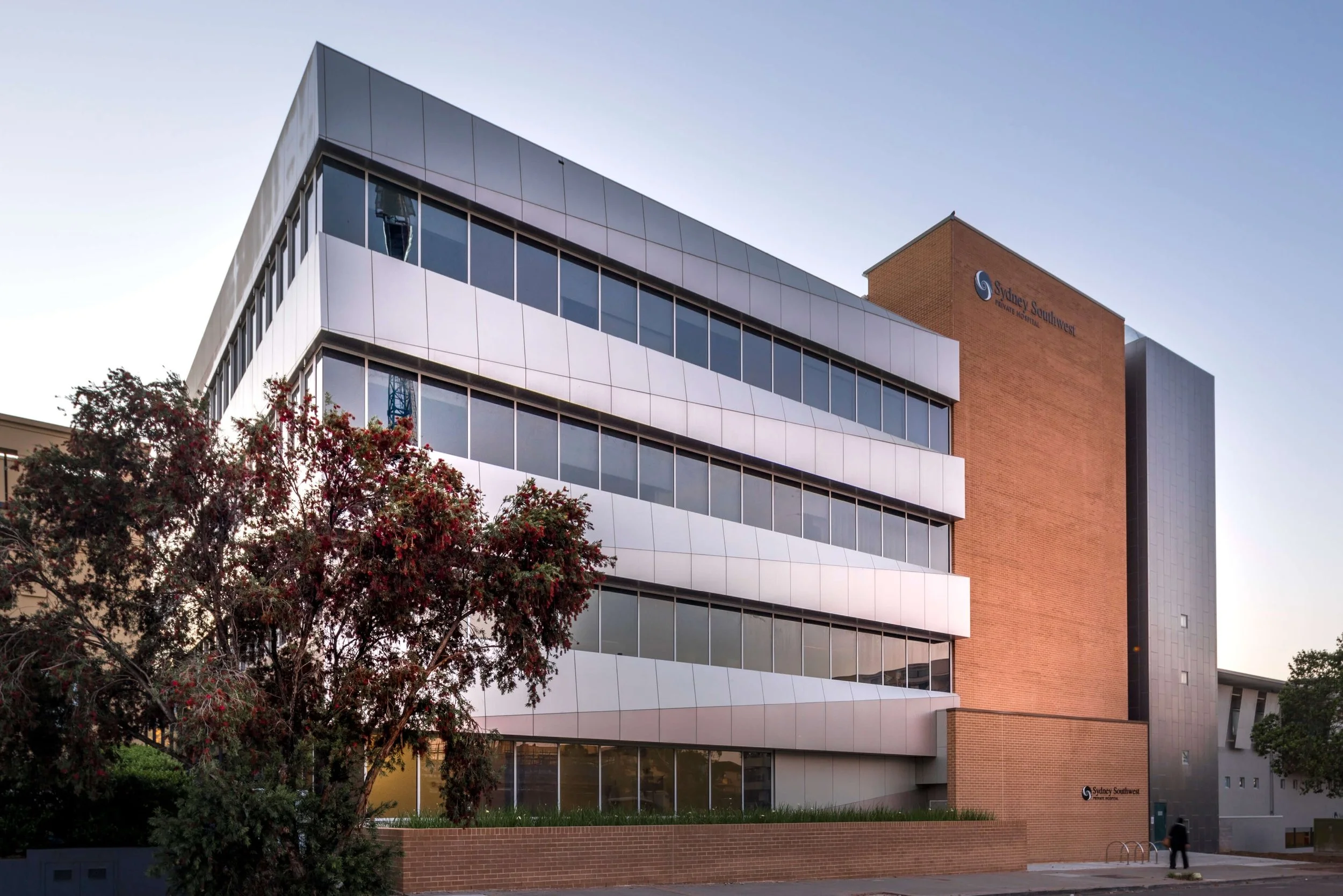 Modern multi-story hospital building with glass windows and brick exterior, with the sign 'Sydney Southwest Private Hospital' on the side, surrounded by trees, under a clear sky.