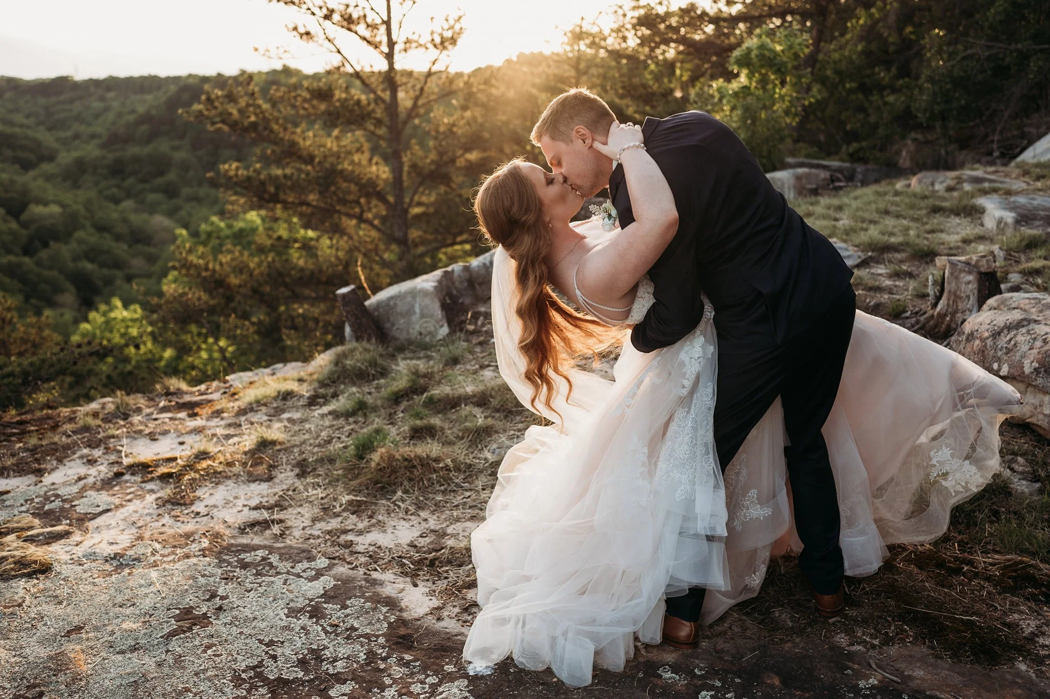 A newlywed couple sharing a kiss on a rocky hilltop at sunset, with trees and a scenic landscape in the background.