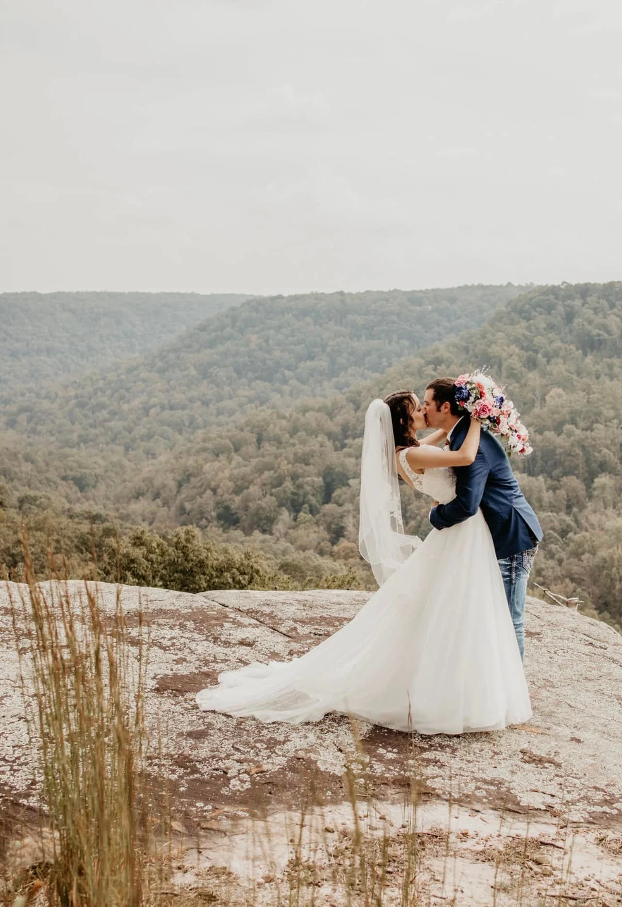 Bride and groom sharing a kiss on a rocky ledge with a scenic backdrop of green mountains and cloudy sky.