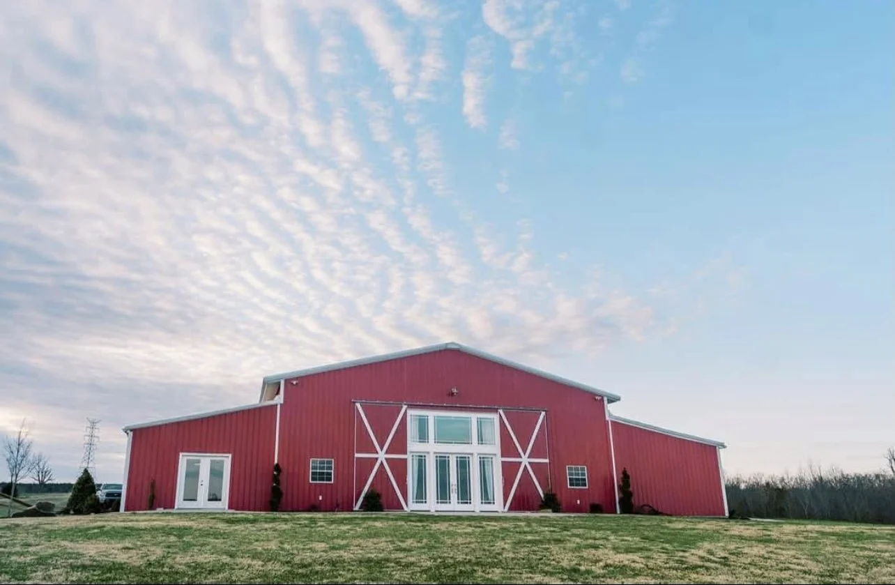 A large red barn with white trim, double doors, and windows, set on a grassy field with a partly cloudy sky above.