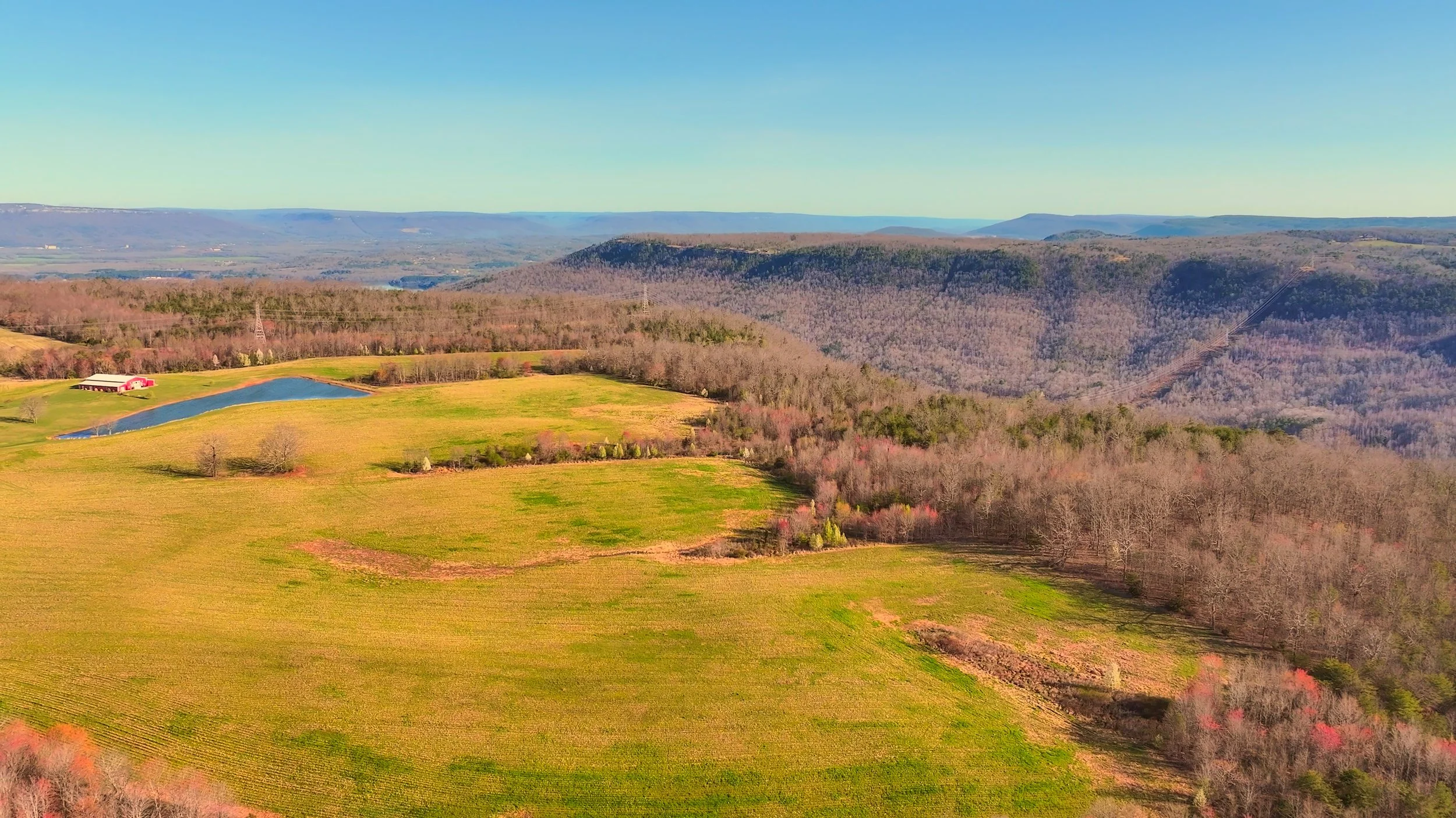 Aerial view of rolling green and brown farmland with a small pond, a building, and forested hills in the distance.