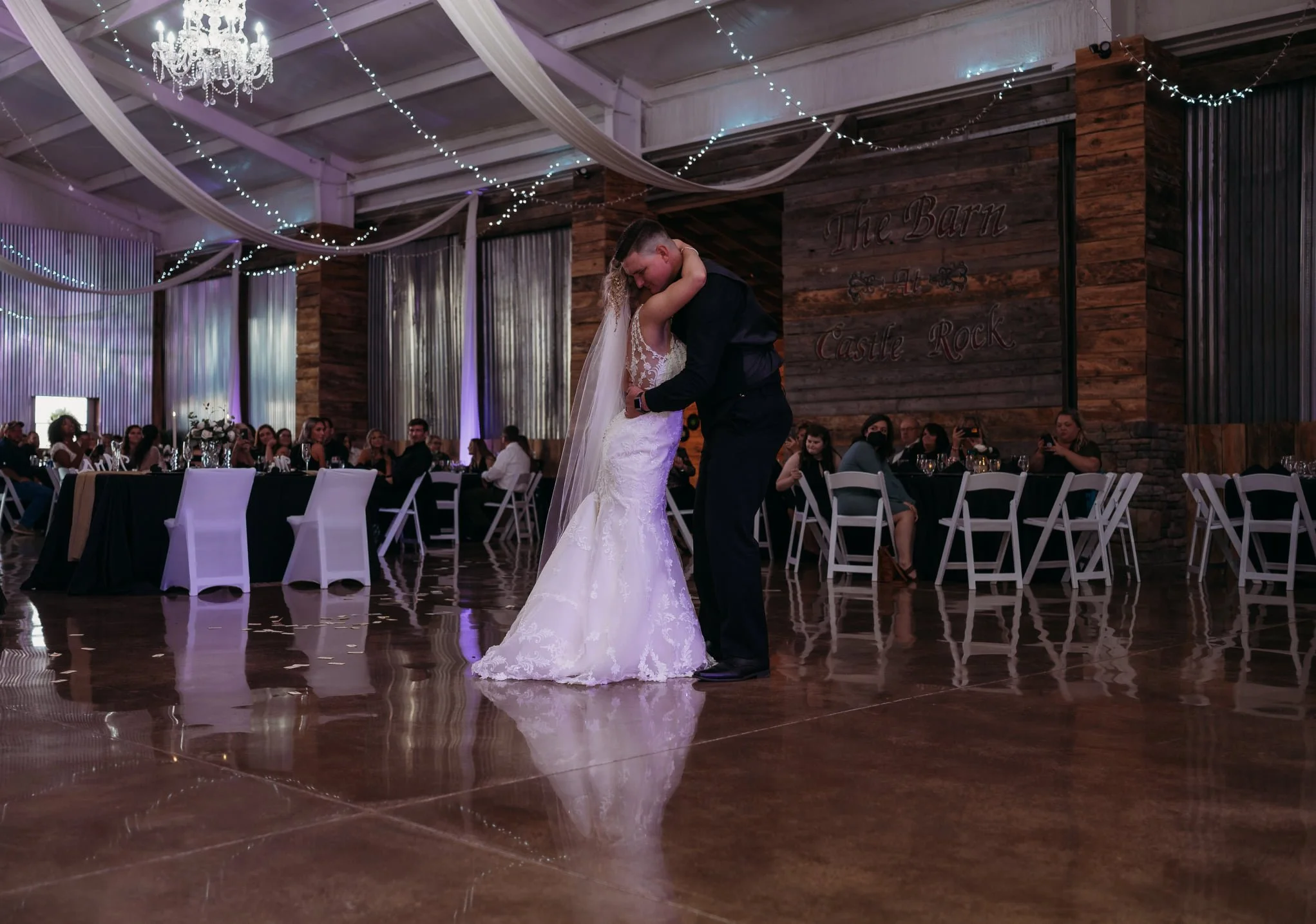 A bride and groom dancing closely at their wedding reception inside a decorated rustic barn with wooden walls and string lights.