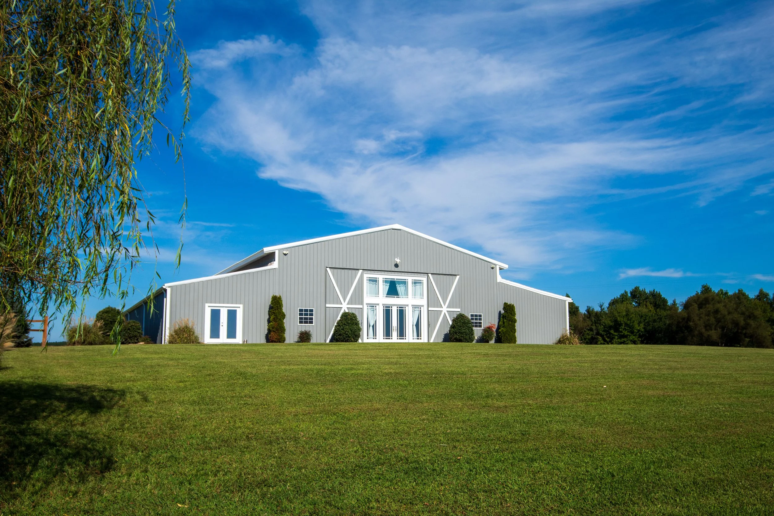 A large light gray barn with white trim and large doors, surrounded by a manicured lawn and small bushes, under a bright blue sky with some wispy clouds.