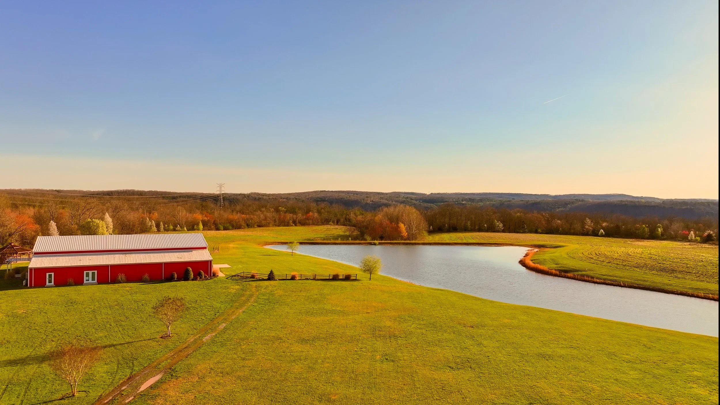 A red barn near a small lake with green grass and trees, under a clear sky, with hills in the background.