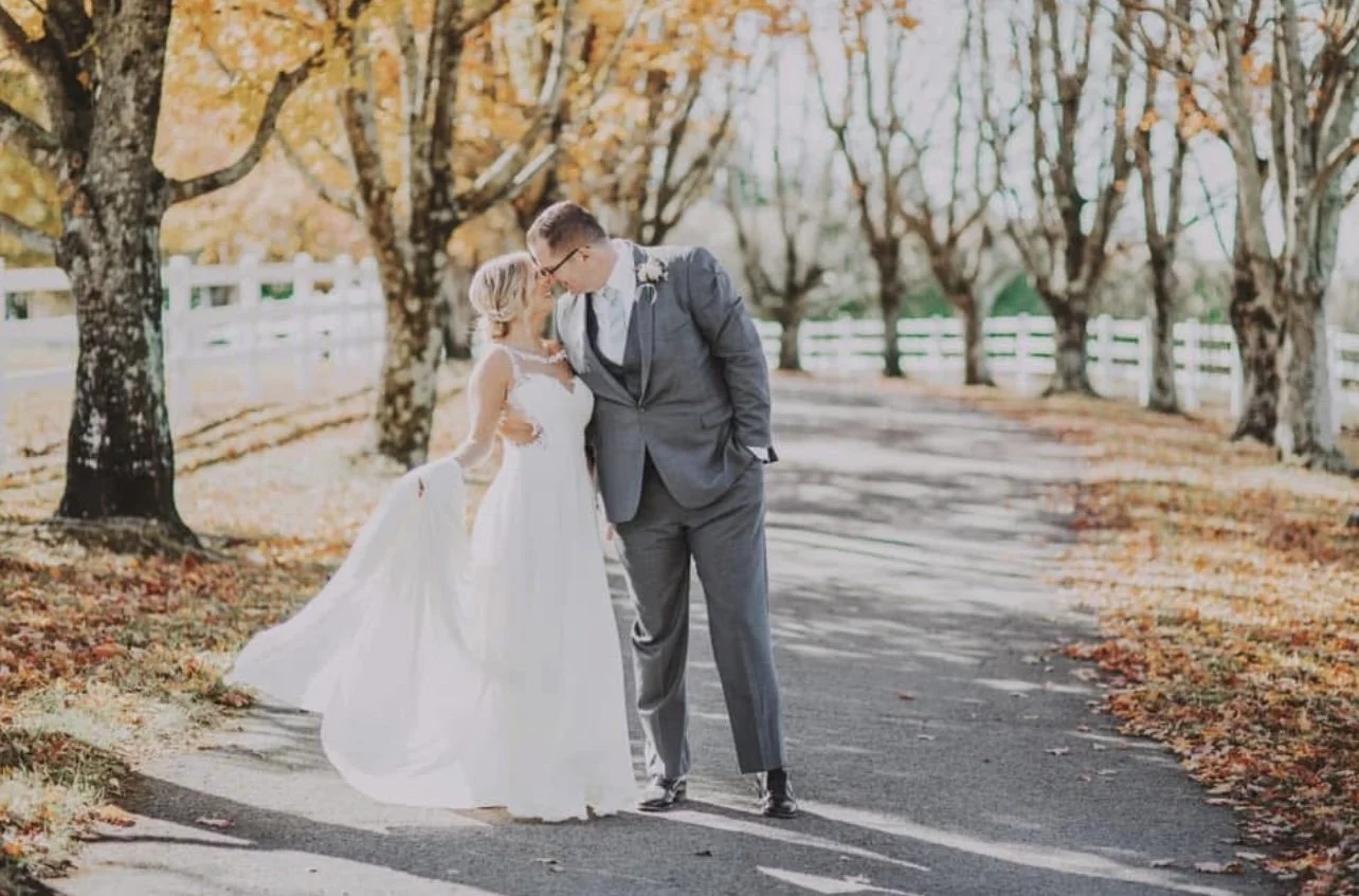 A bride and groom dressed in wedding attire sharing a tender moment outdoors on a tree-lined path with fall foliage.