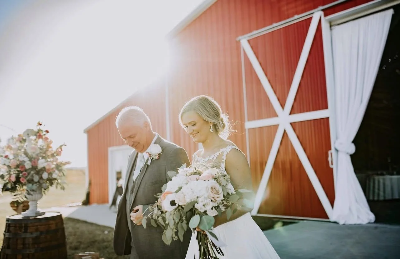 A bride and an older man, likely her father, standing outdoors during a wedding ceremony. The bride is holding a bouquet of white and pink flowers, wearing a white wedding dress with lace details, and smiling. The background features a red barn with white accents, and the scene is bathed in sunlight.