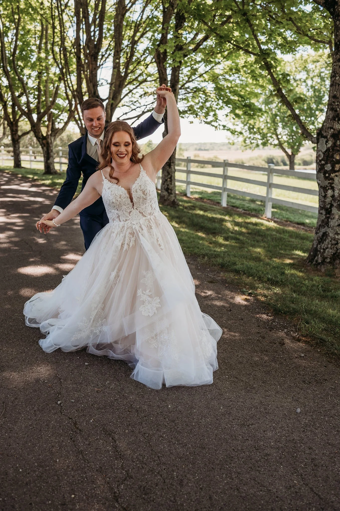 A bride and groom dancing outdoors on a tree-lined path, with the groom twirling the bride in her wedding gown.