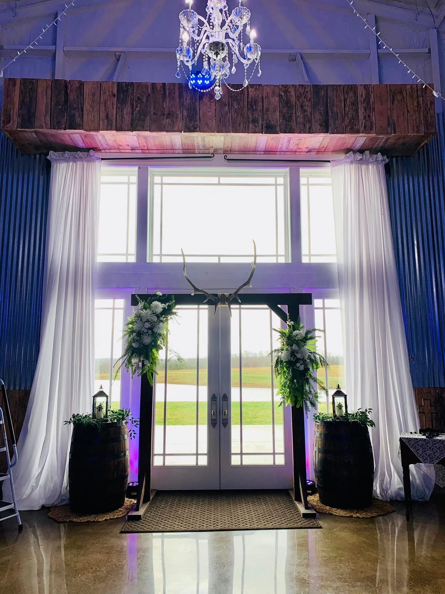 Elegant entrance with white drapes, floral arrangements, lanterns, and antlers above double glass doors, with a chandelier hanging from the ceiling.