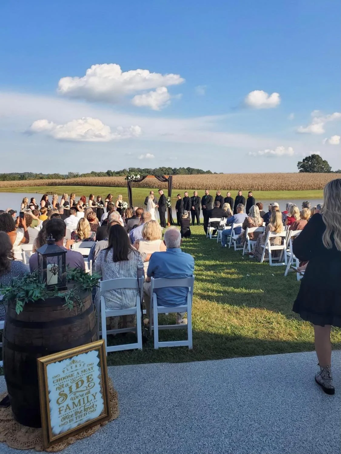 Outdoor wedding ceremony with guests seated on white chairs, officiant and couple at the altar, set against farmland and a blue sky with clouds.