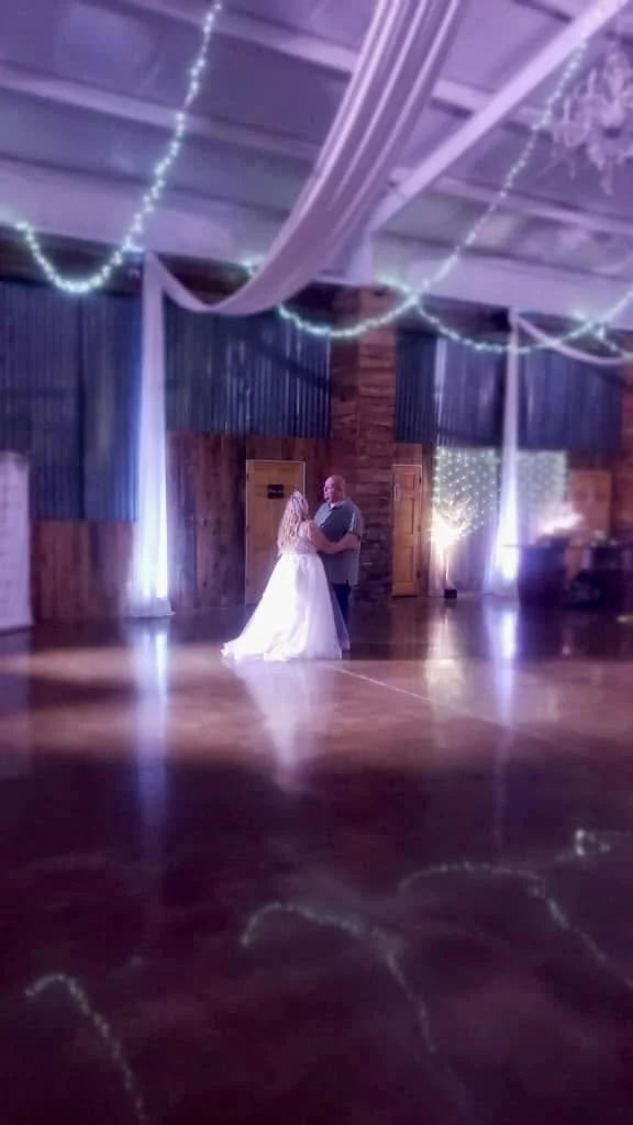 A bride and groom sharing their first dance at a wedding reception in a decorated barn, with purple and white lighting and string lights overhead.