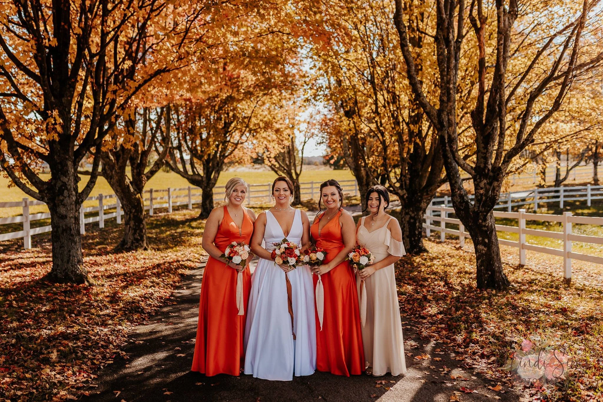 Four women in dresses posing on a tree-lined path with fall foliage, holding bouquets of flowers