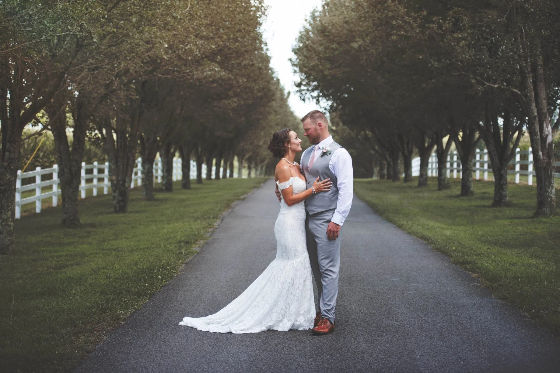 A bride and groom standing close together on a tree-lined pathway, gazing into each other's eyes, with the bride wearing a white lace wedding dress and the groom in a gray vest, white shirt, and gray pants.