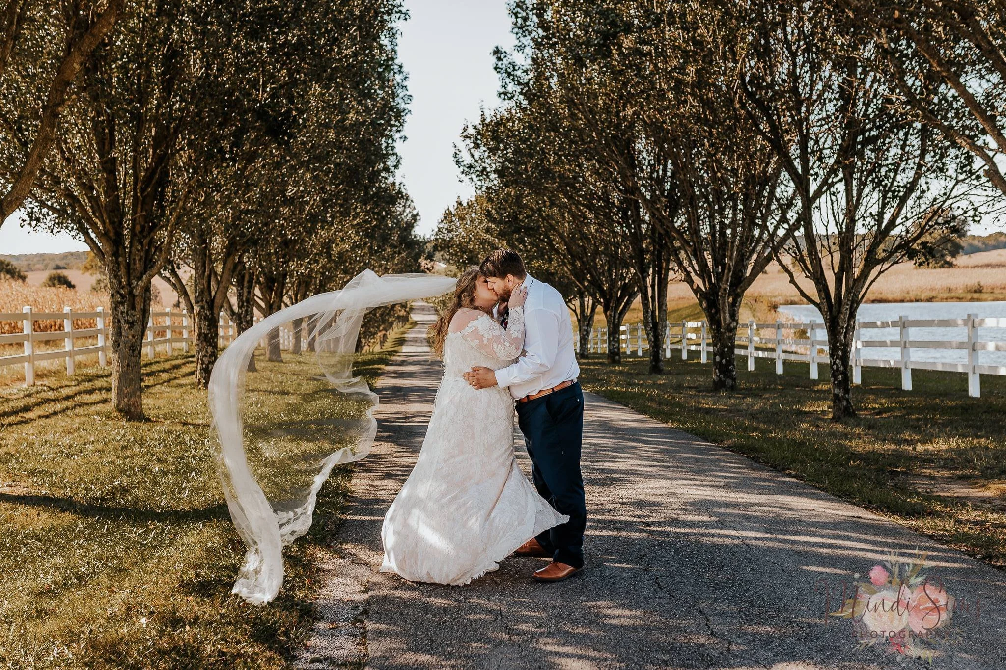 A bride and groom kissing on a tree-lined country road during sunset, with the bride in a white lace wedding dress and the groom in a white shirt and dark pants.