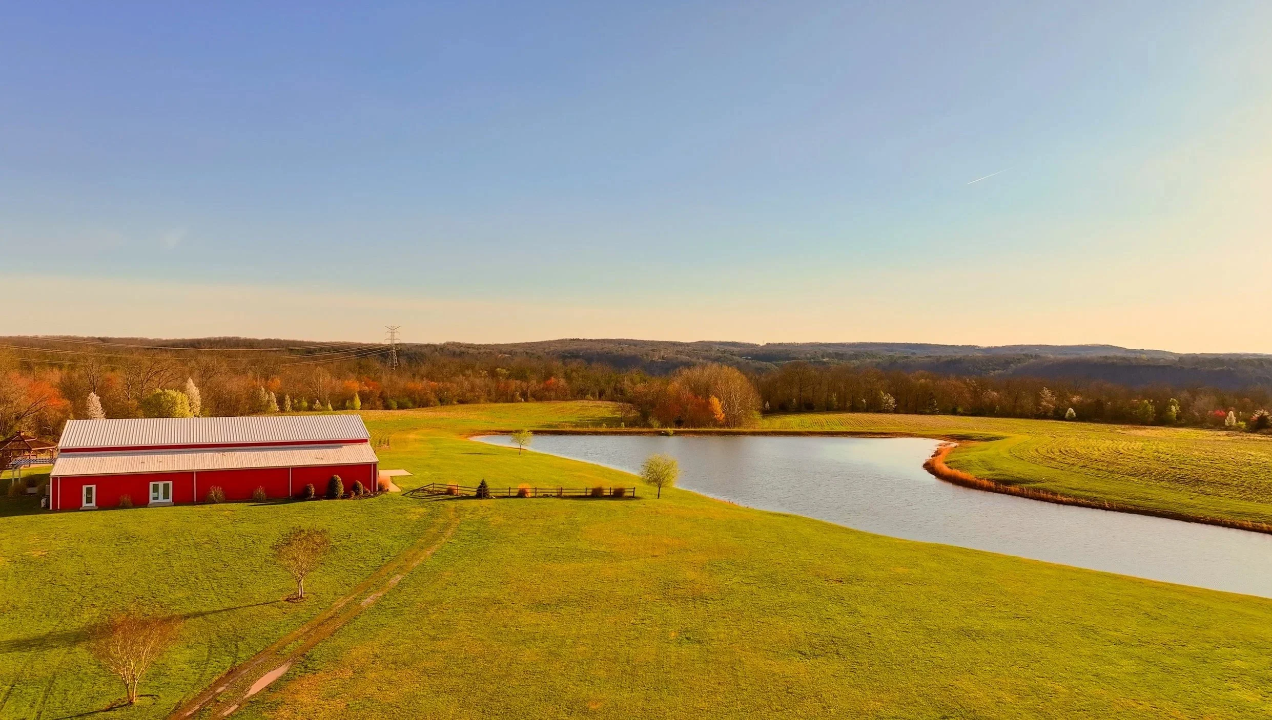 A rural landscape with a red barn, a pond, green fields, and trees with autumn colors under a clear blue sky.
