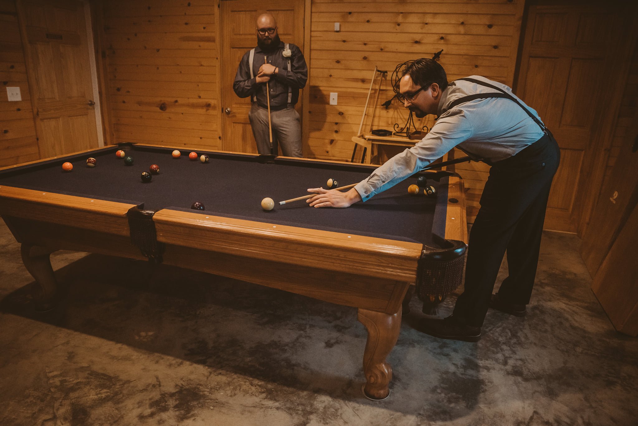Two men playing pool in a wooden-paneled game room, one taking a shot and the other observing with a cue stick in hand.