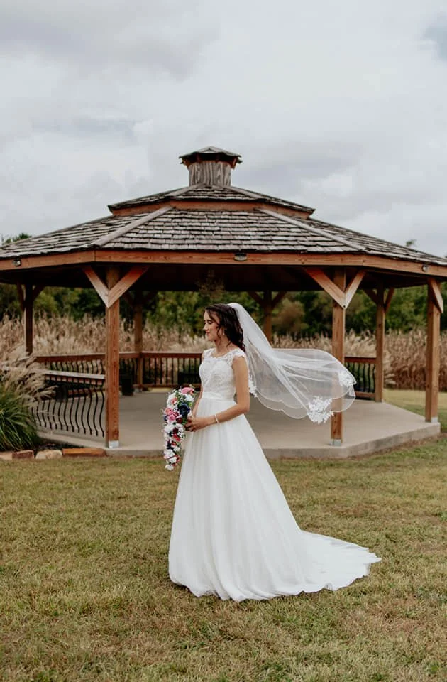 Bride in a white wedding dress and veil holding a floral bouquet, standing outdoors in front of a wooden gazebo with a cloudy sky overhead.