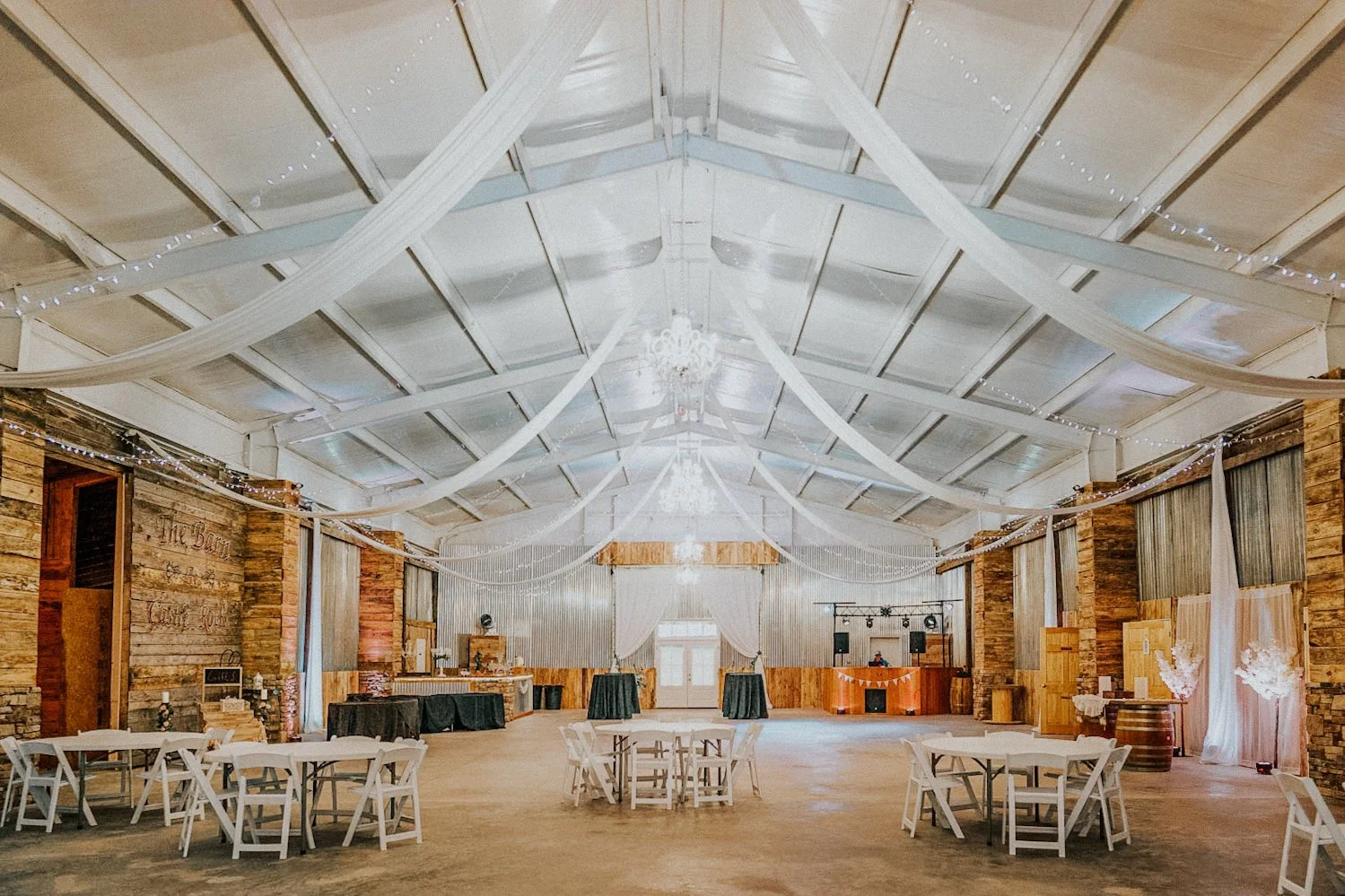 Interior of a rustic wedding barn decorated for a celebration, with white drapes hanging from the ceiling, string lights, chandeliers, round tables with white chairs, and a DJ booth at the back.