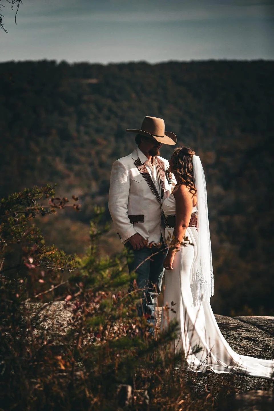 A couple dressed in wedding attire standing close together outdoors with a mountain landscape in the background.
